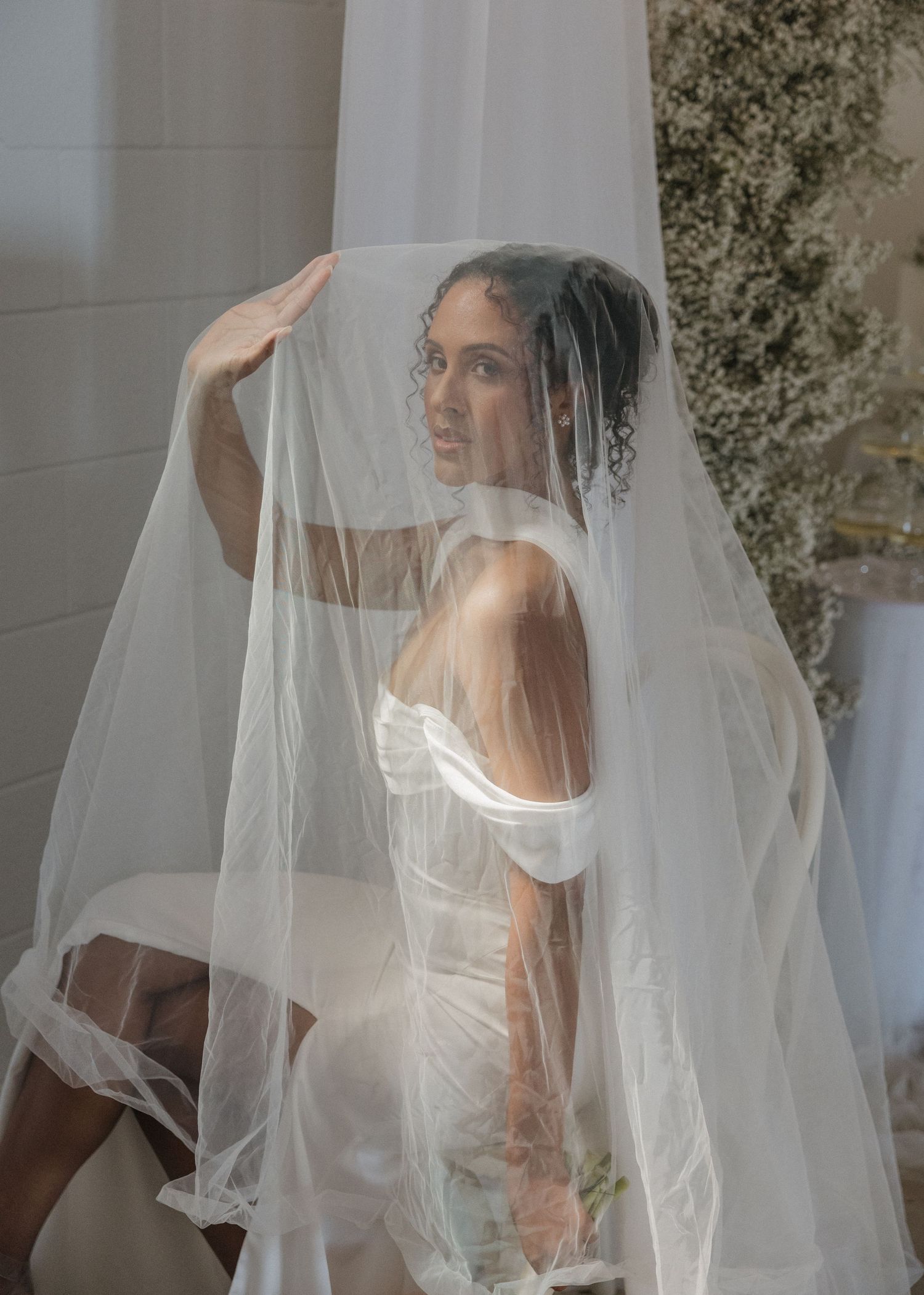 Bride in off-shoulder gown draped under flowing white veil, posing elegantly in a softly lit bridal setting.