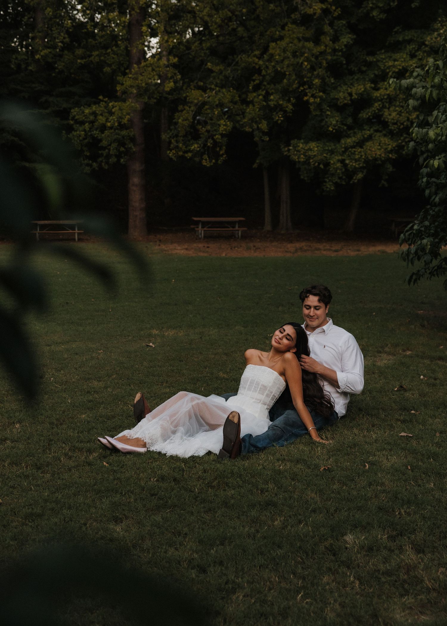 Couple sitting on green grass in a park, woman in white dress leaning against man in white shirt and jeans.