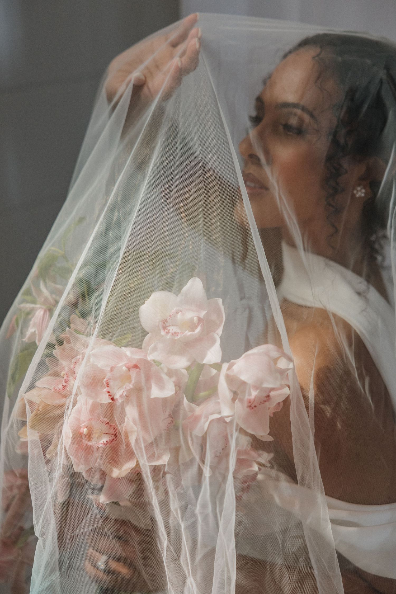 Black bride in white dress holds pink orchids under sheer veil in elegant bridal portrait.