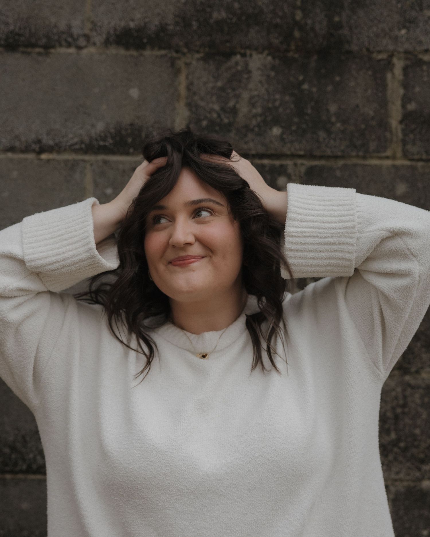 A young woman in a cream sweater smiles upward, hands in dark wavy hair, posed against a stone wall.