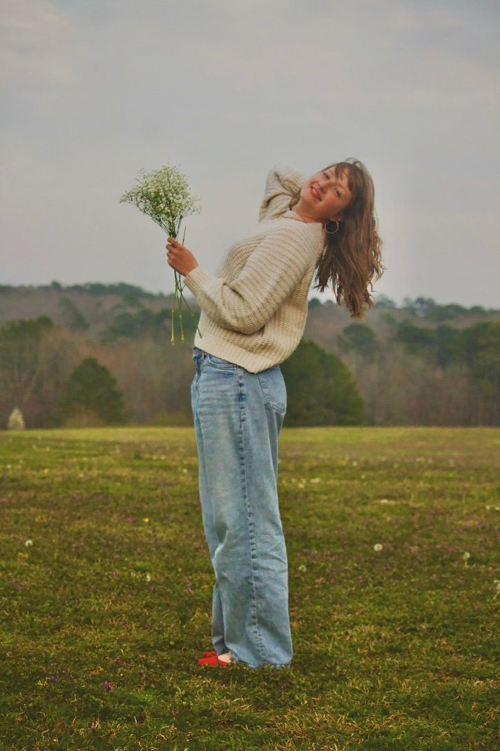 Casual pose in sweater and jeans while holding flowers in open field.