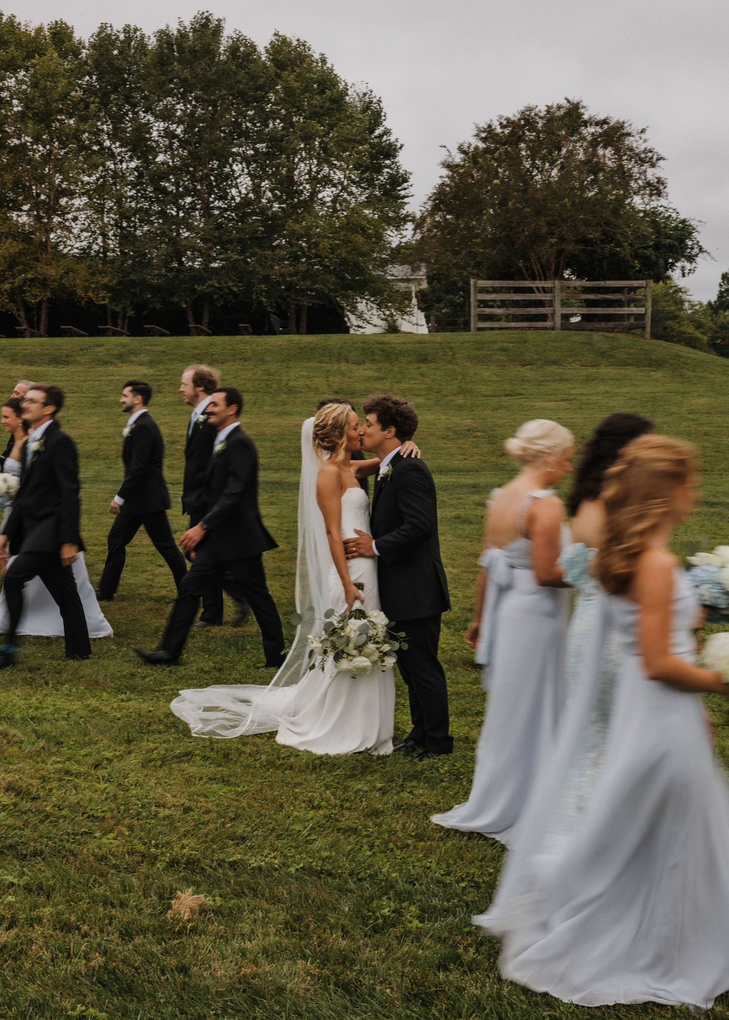 Bride and groom share a kiss surrounded by wedding party in black suits and light blue bridesmaid dresses on a green lawn.