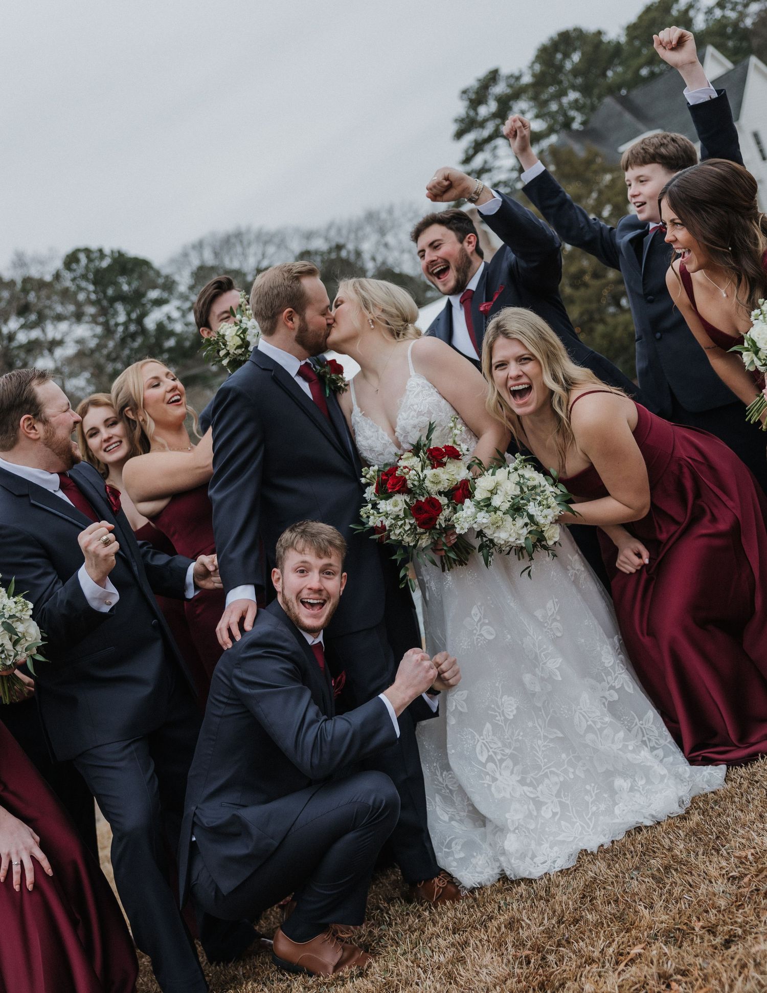 Joyful wedding party in navy and burgundy celebrating with bride and groom kissing outdoors.