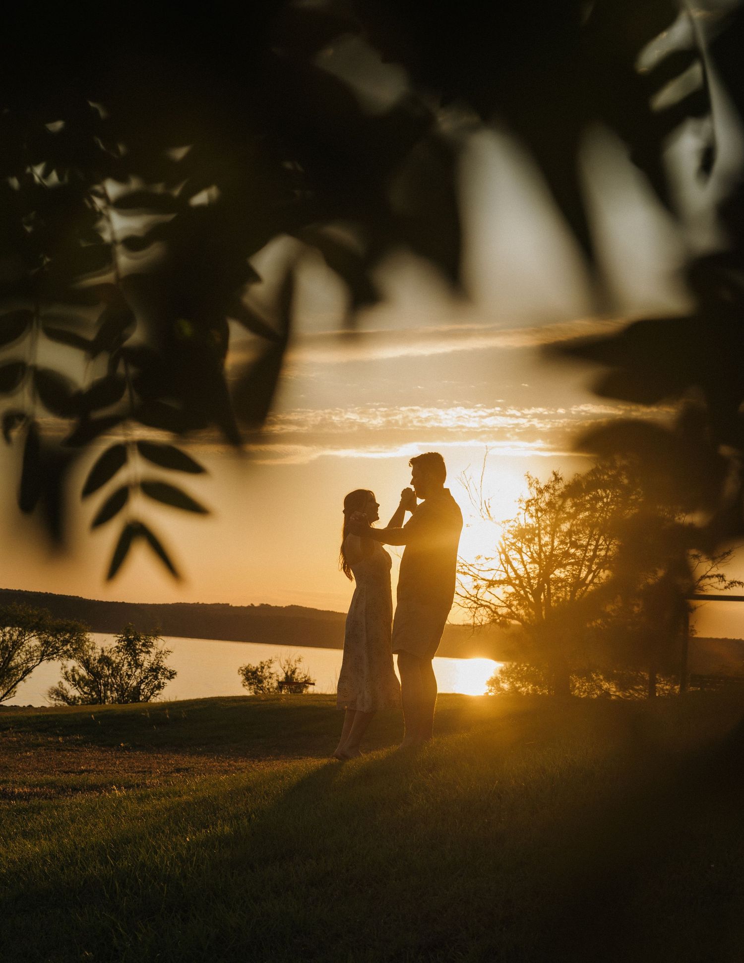 Couple silhouetted against golden sunset by a lake, framed by leaves, sharing a romantic moment.