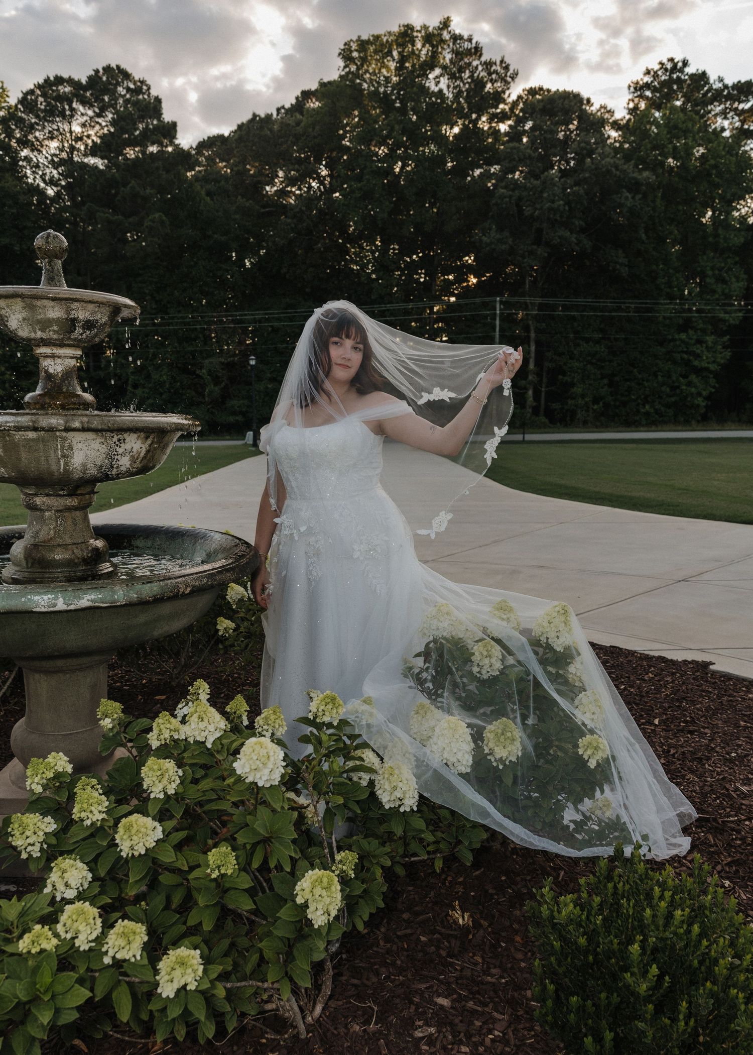 Bride in white off-shoulder gown and flowing veil poses by stone fountain with white hydrangeas at dusk.