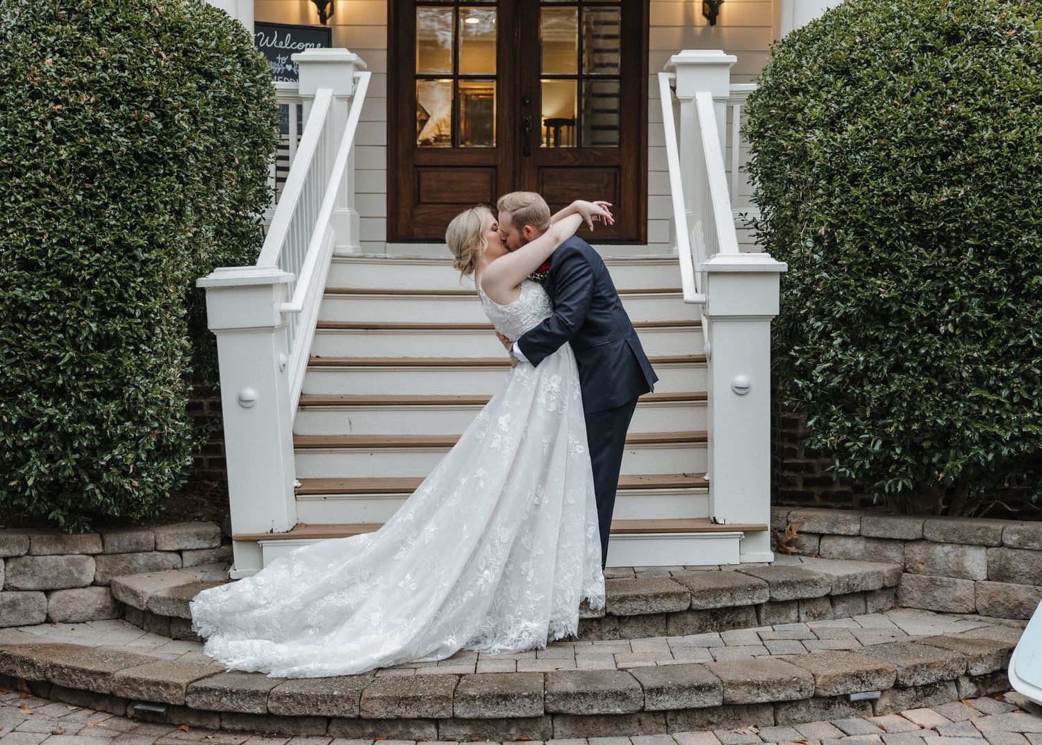 Bride and groom share romantic embrace on elegant white staircase surrounded by lush greenery at wedding venue.