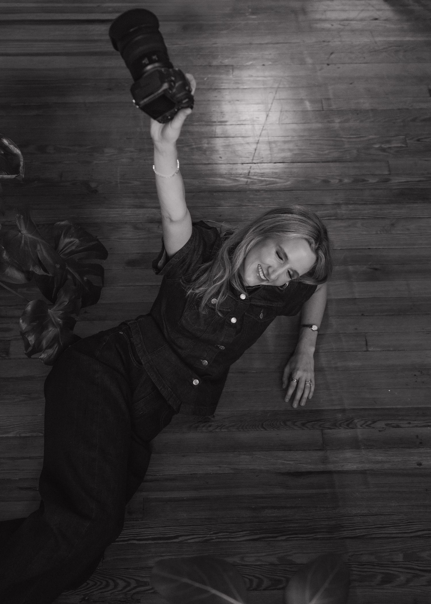 Black and white photo of a blonde woman lying on hardwood floor holding a DSLR camera up in the air.