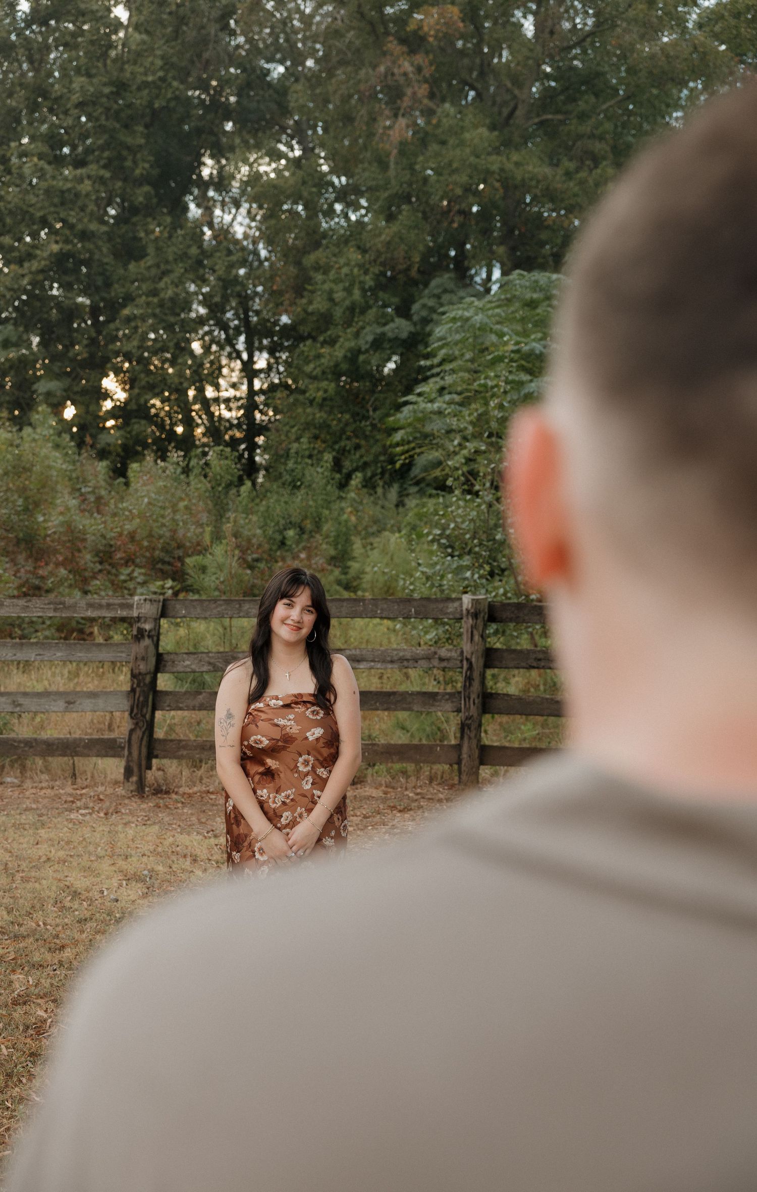 A couple shares an intimate moment in a rustic outdoor setting with wooden fences and trees in the background.