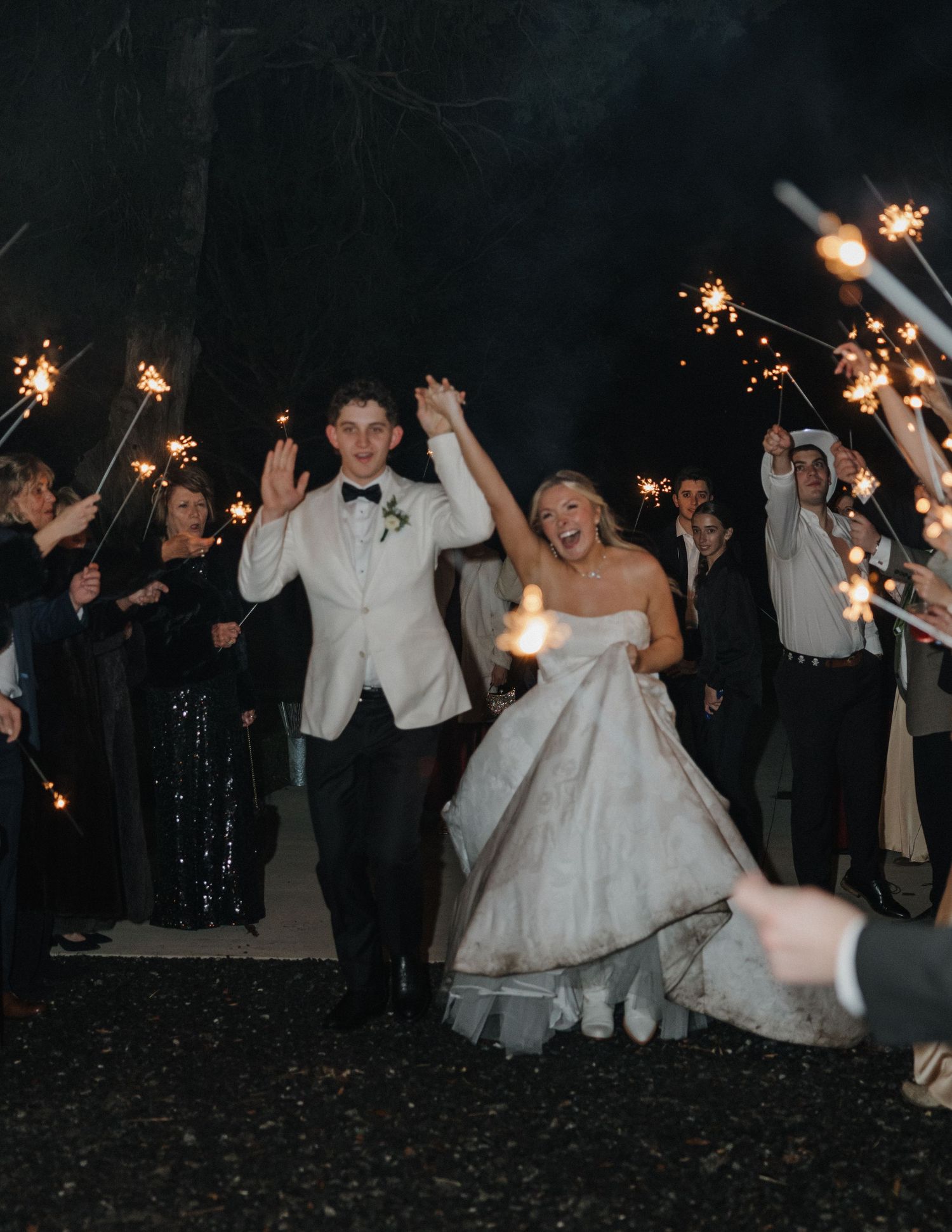 Wedding party celebrates at night with sparklers while dancing and posing for photos on a dark outdoor patio.