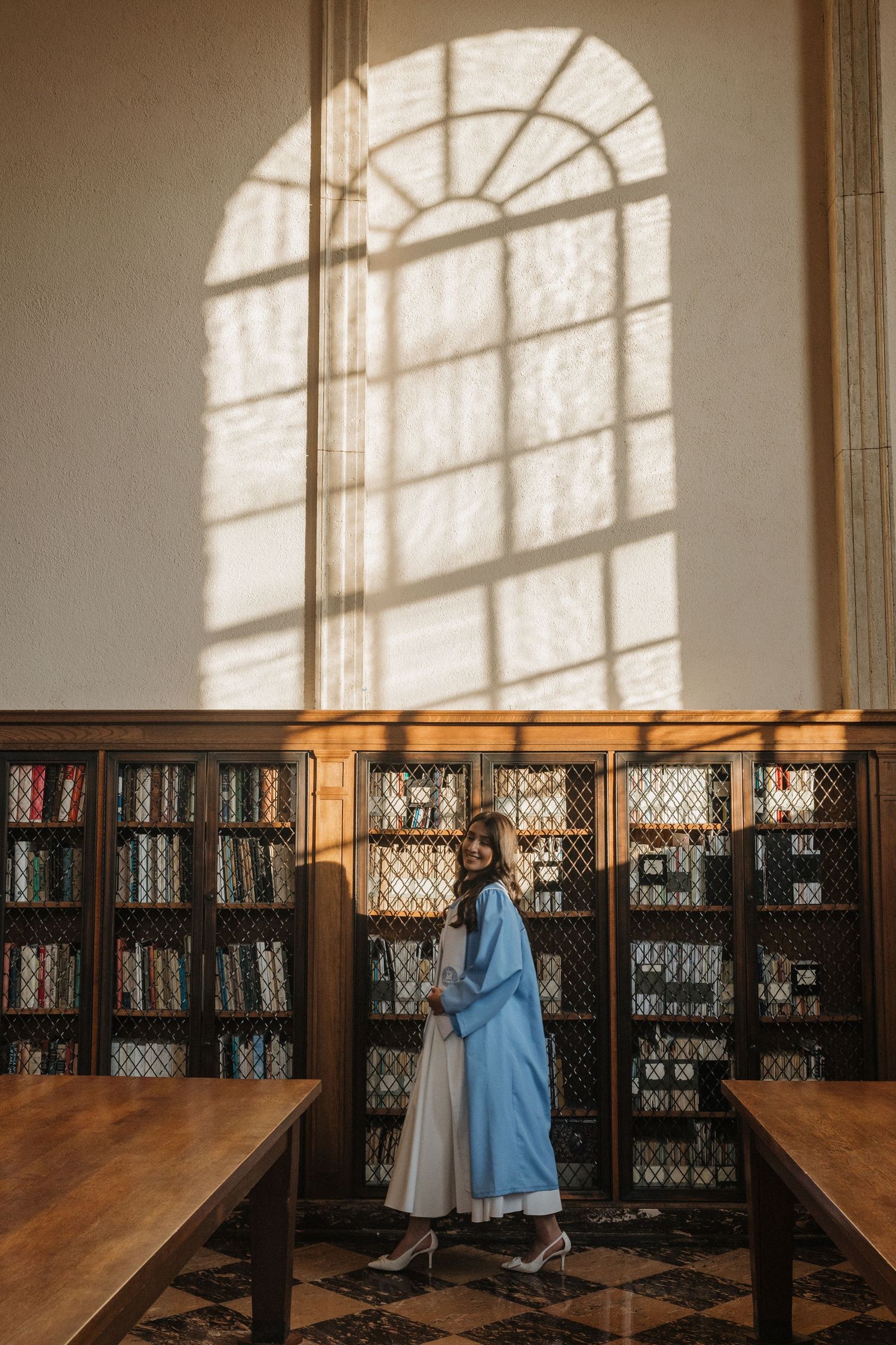Woman in blue graduation gown standing in sunlit library with wooden bookshelves and arched window.