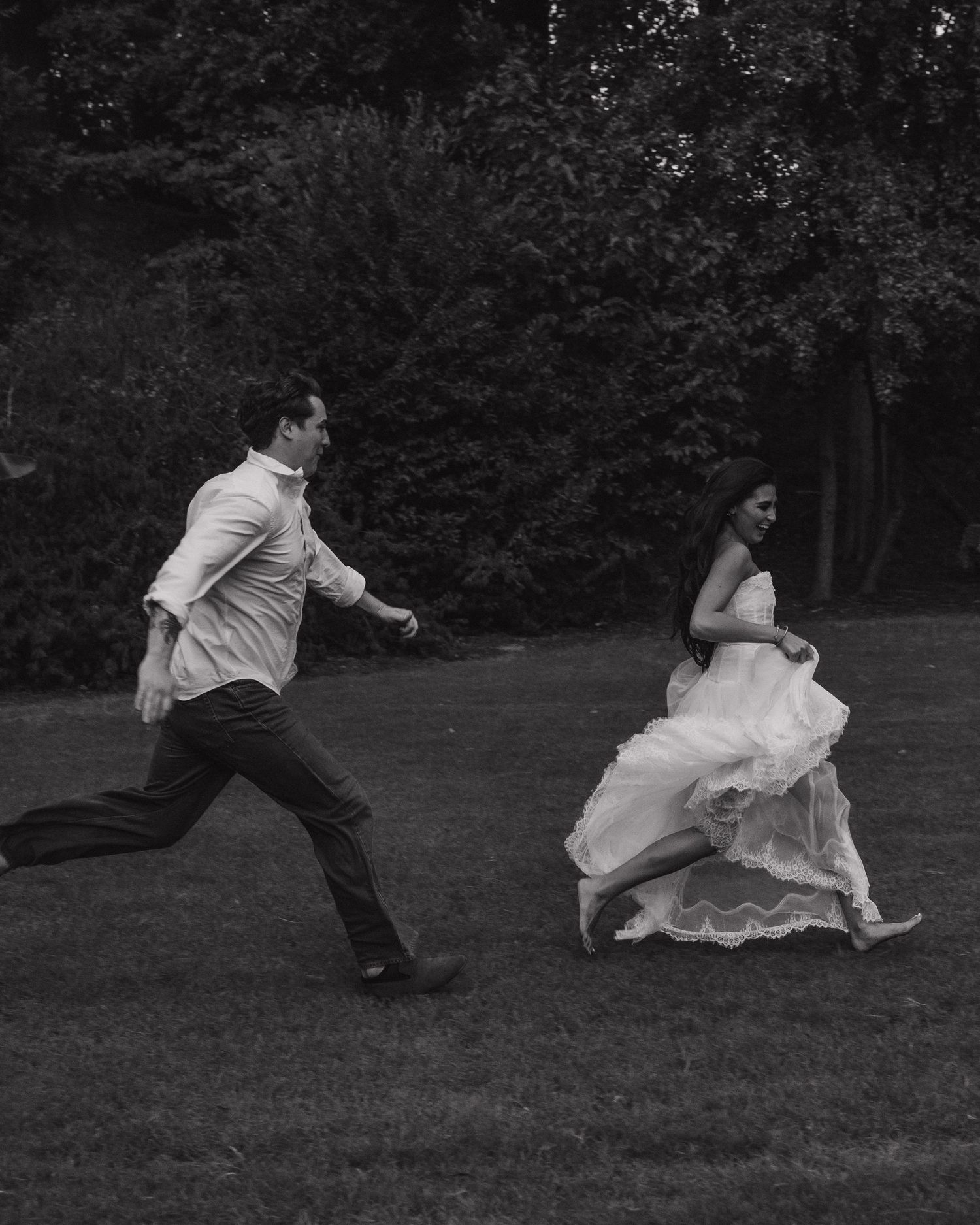 Black and white photo of a bride in a flowing gown and groom in white shirt running playfully across a lush green lawn.