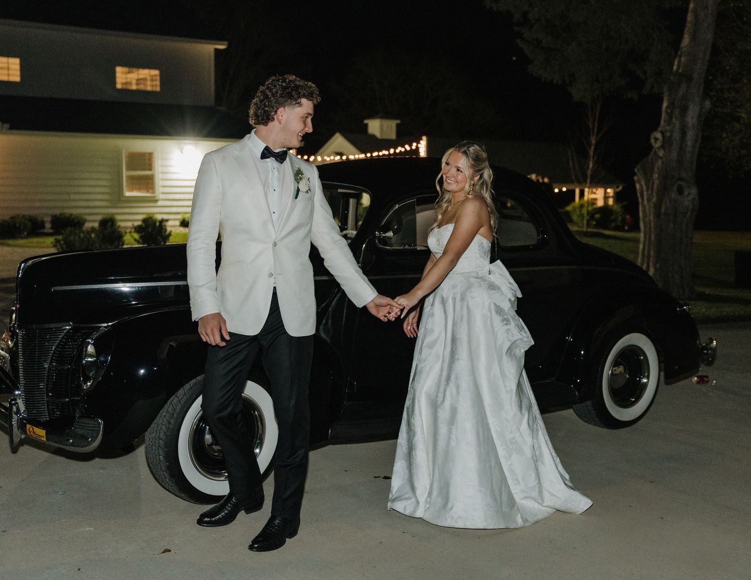 Bride and groom holding hands beside a vintage black car at night, groom in white tuxedo, bride in elegant white gown.
