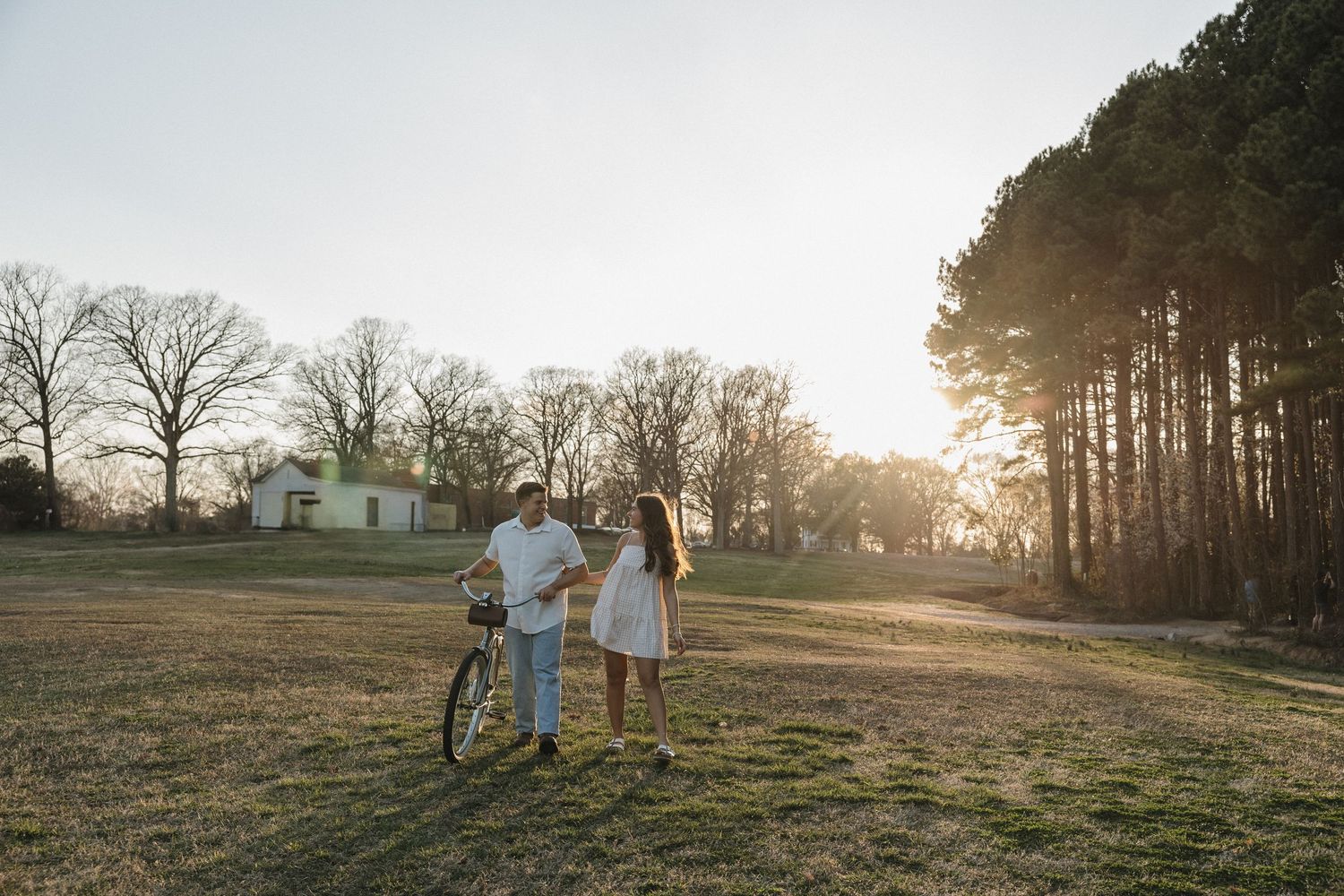 Couple standing together in a sunlit field with a bicycle, tall trees and a white house in the background.