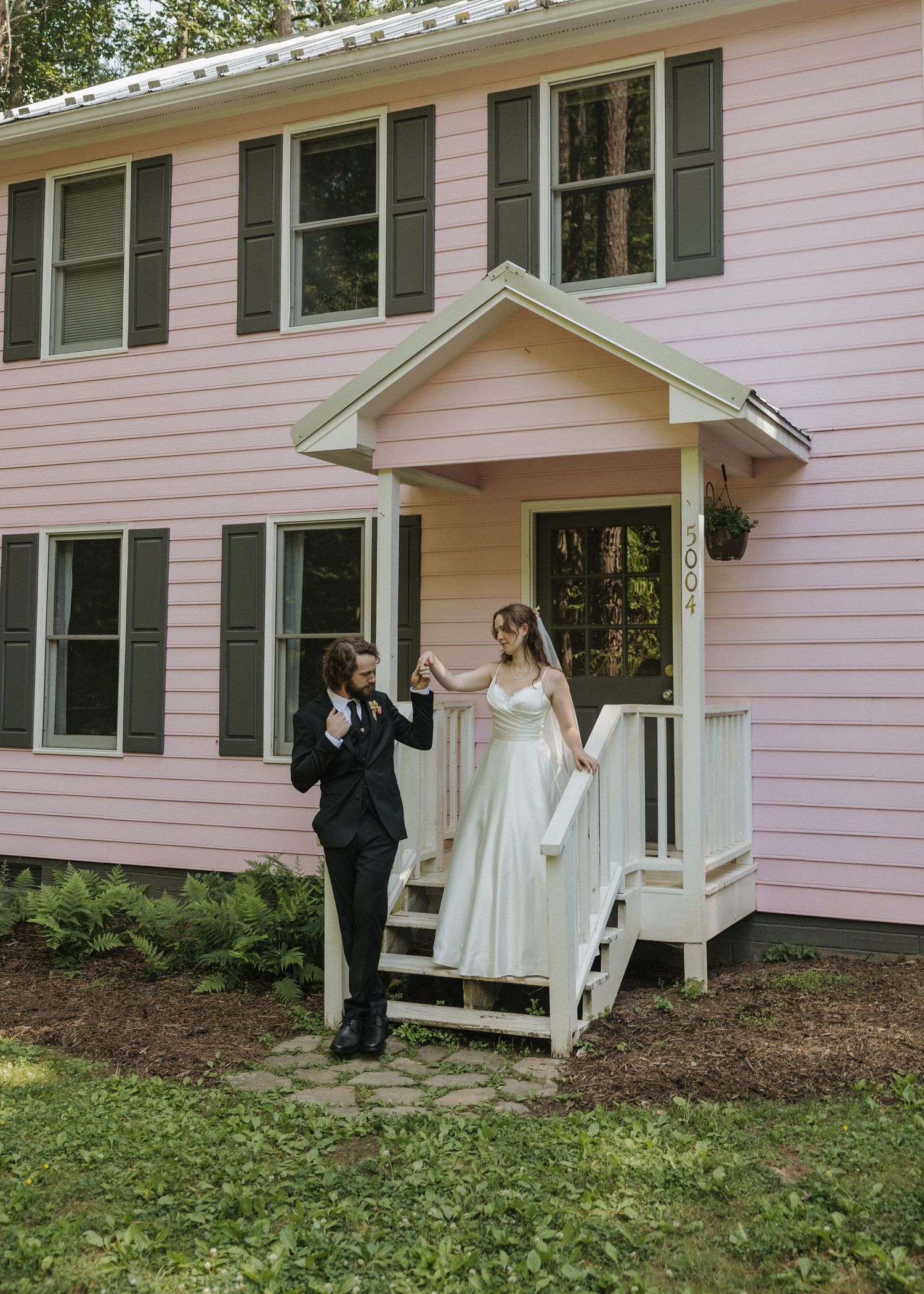 Bride and groom in wedding attire holding hands on steps of charming pink colonial house with dark shutters.