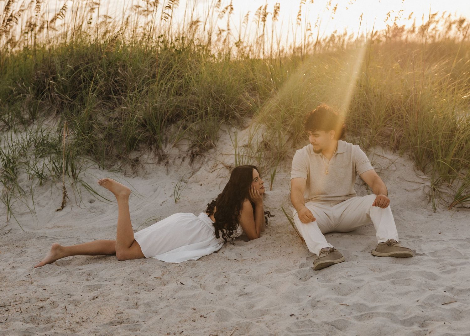 Couple sitting and lying on sandy beach dunes at golden hour with tall grass in warm sunset light.
