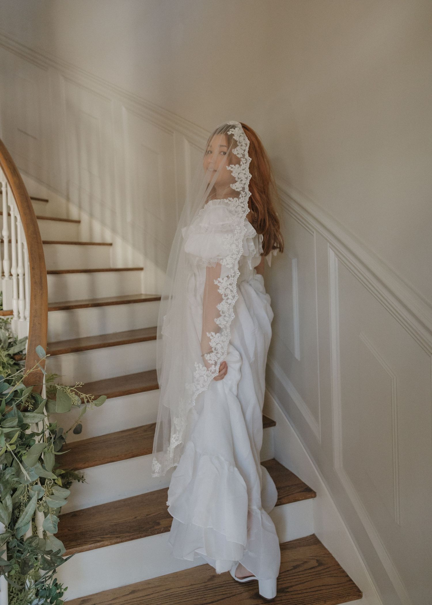 A bride in a flowing white lace dress with a long veil ascends a wooden staircase decorated with greenery.