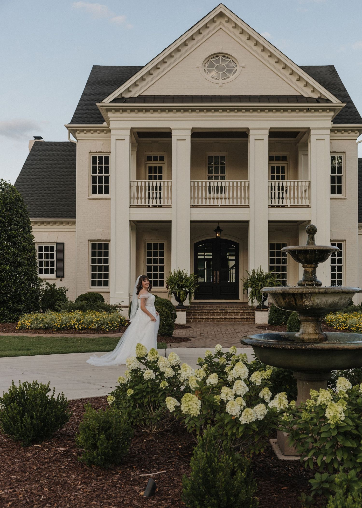 Bride in white gown poses by fountain outside elegant Southern colonial mansion with white columns and manicured gardens.