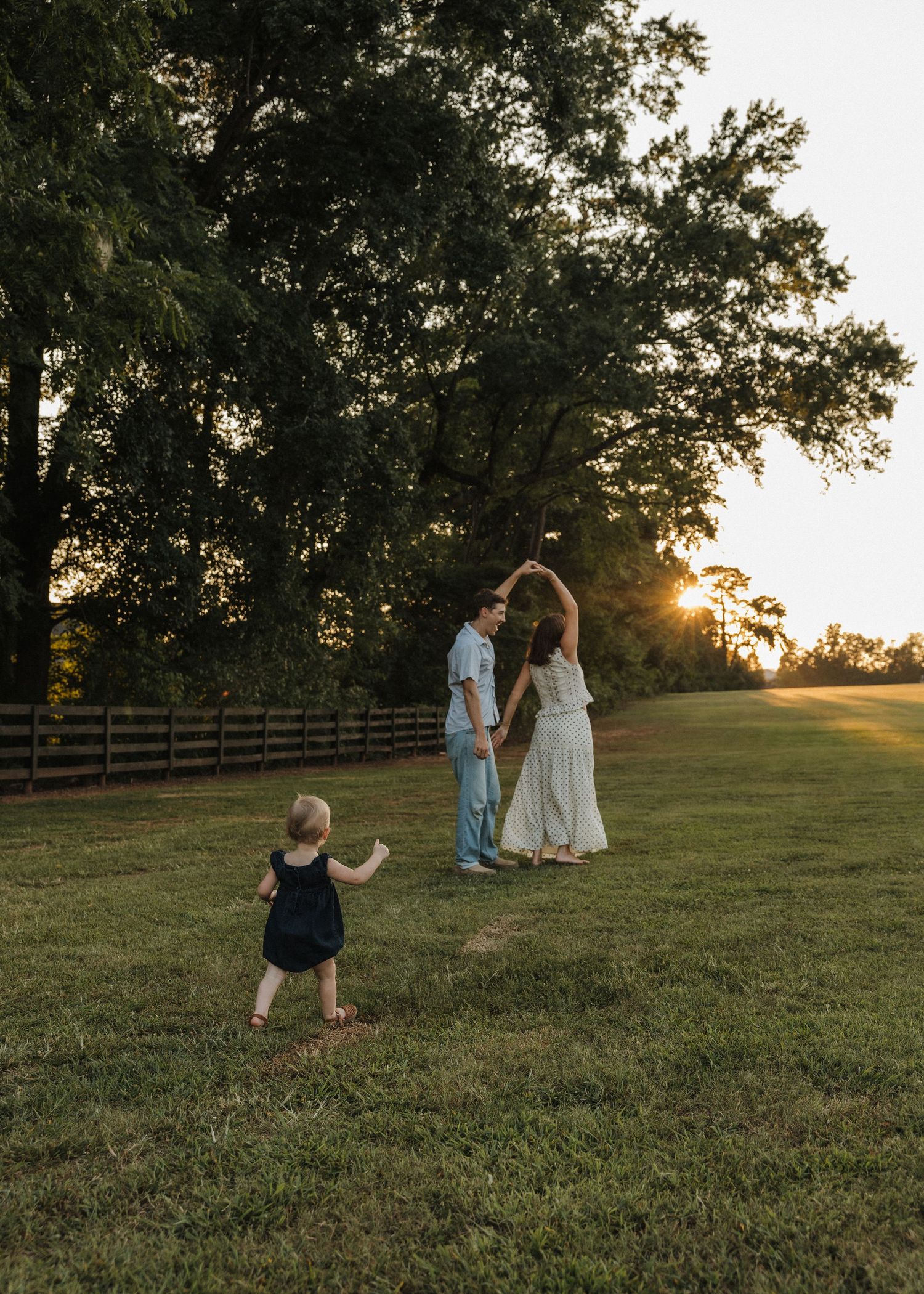 Family dancing at golden hour in a green field near a wooden fence with large trees at sunset.