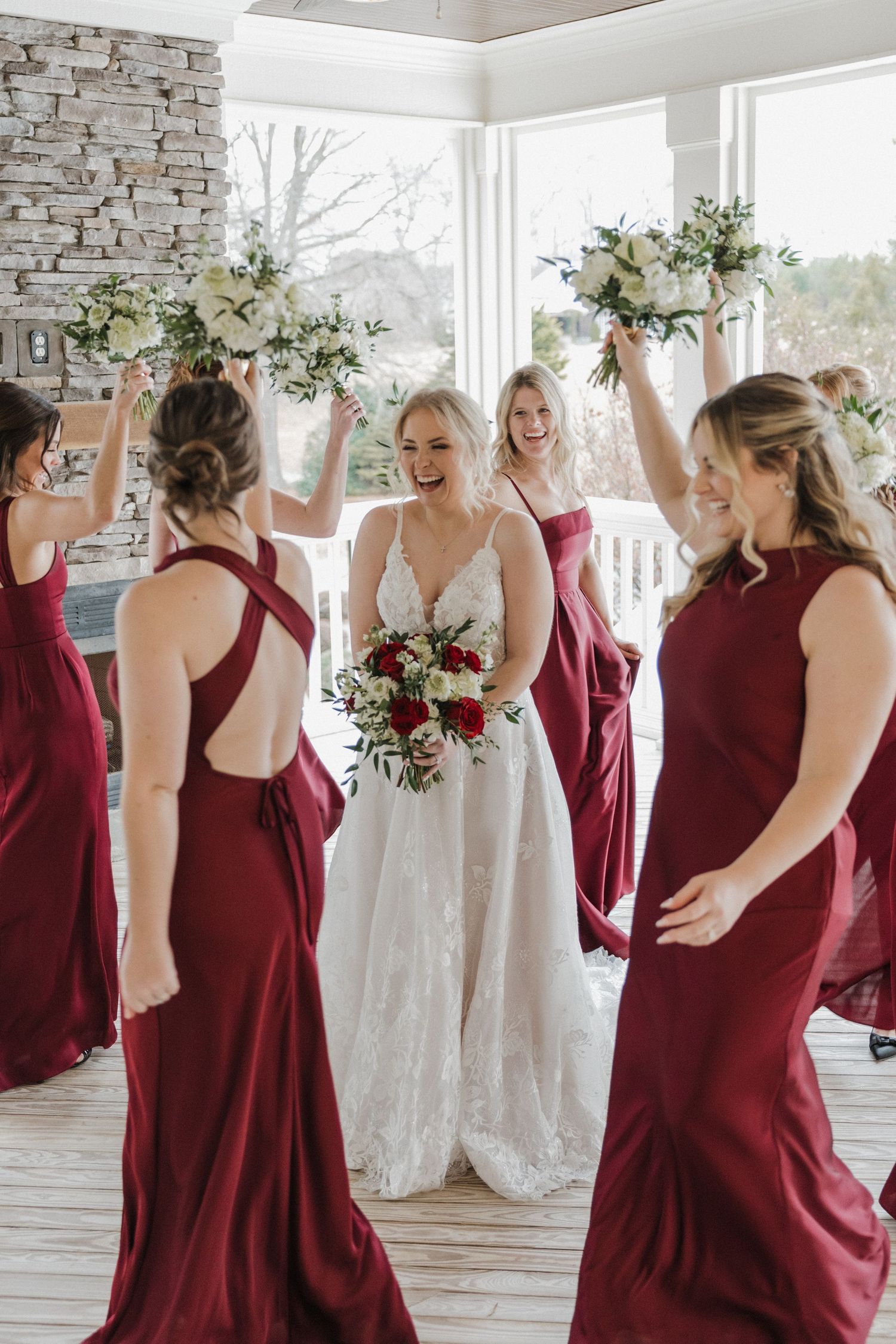 Bride in white lace gown surrounded by bridesmaids in burgundy dresses raising bouquets in celebration indoors.