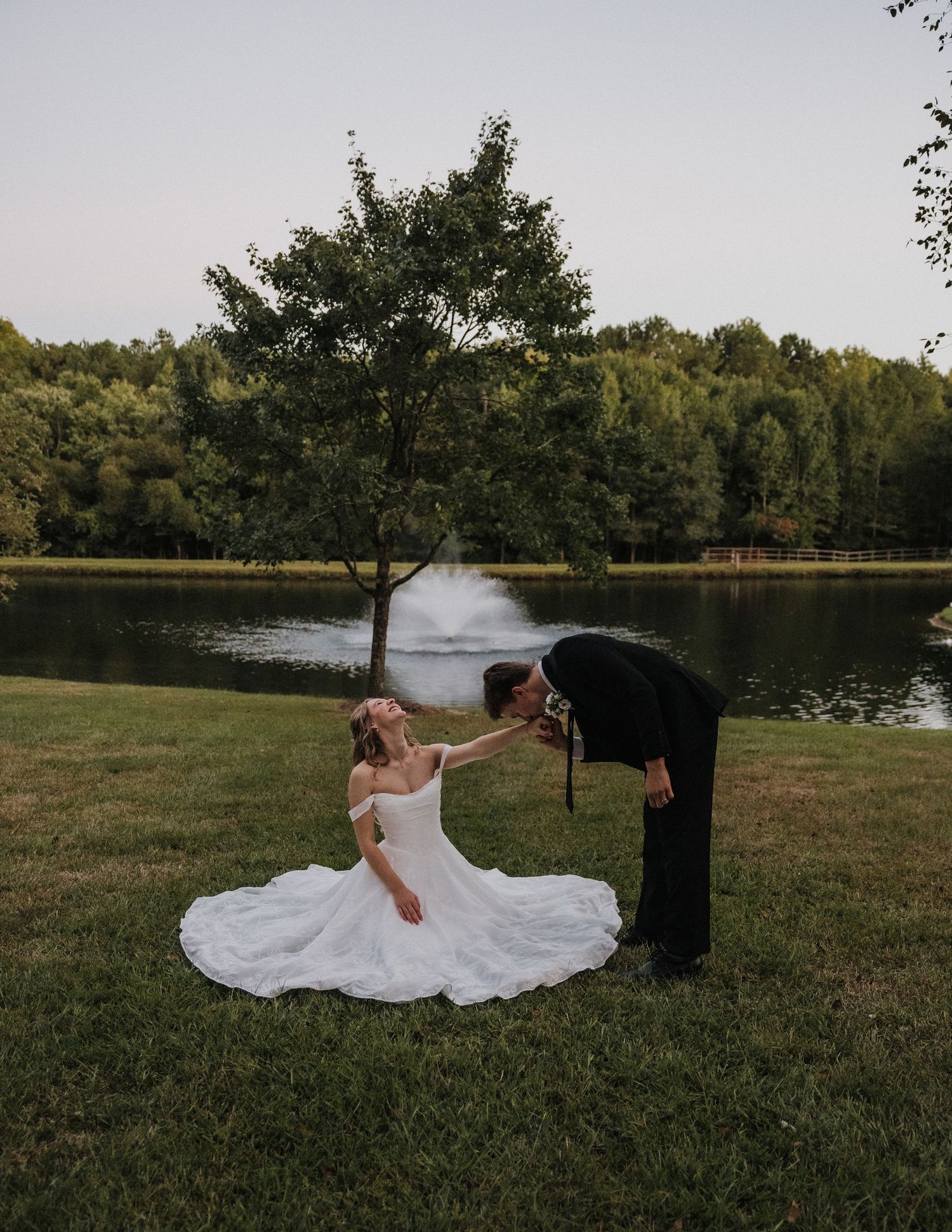 Bride in white gown sits on grass as groom in black suit kisses her hand by a serene pond with fountain.