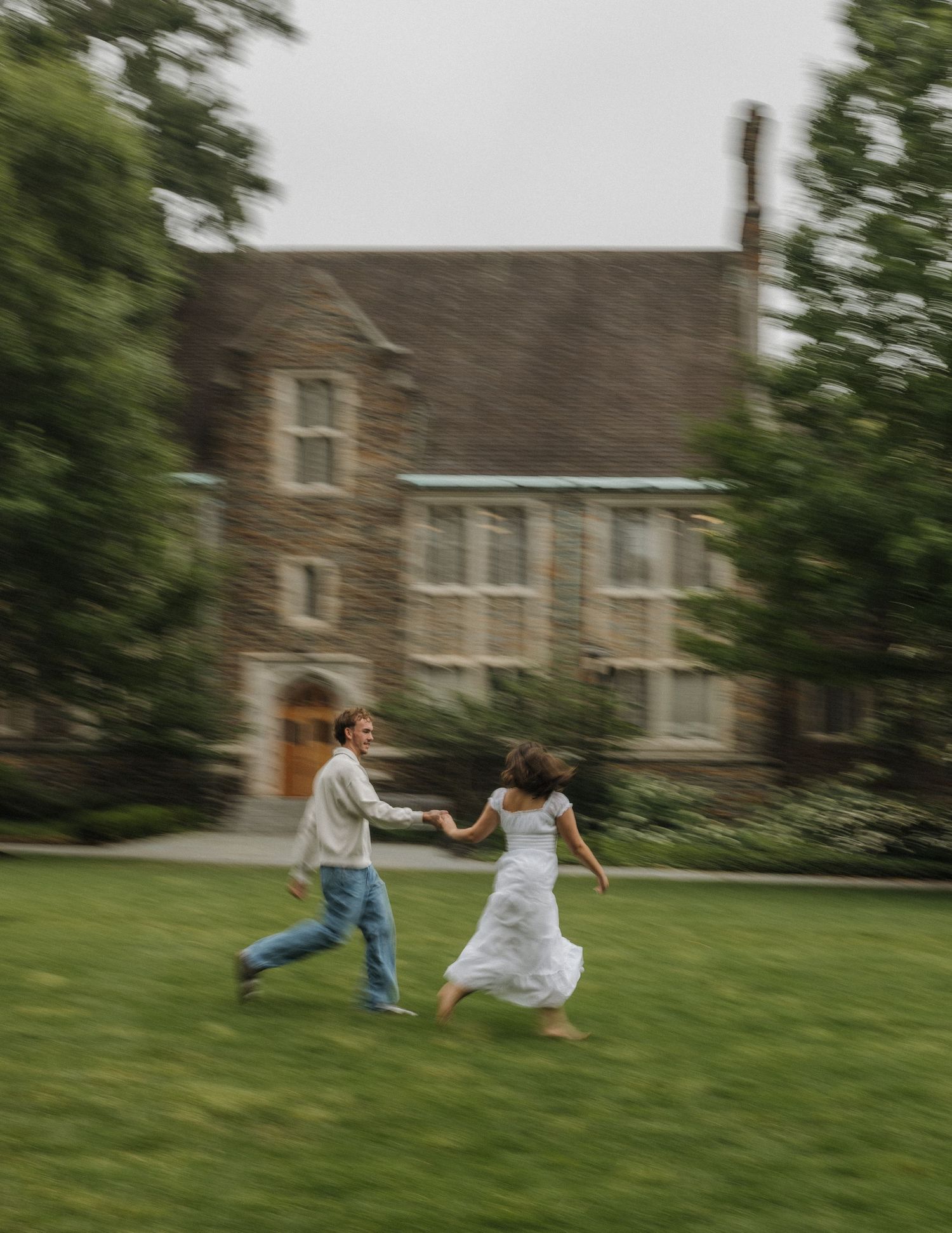 A couple runs hand-in-hand across a green lawn in front of a stone building, captured with motion blur.