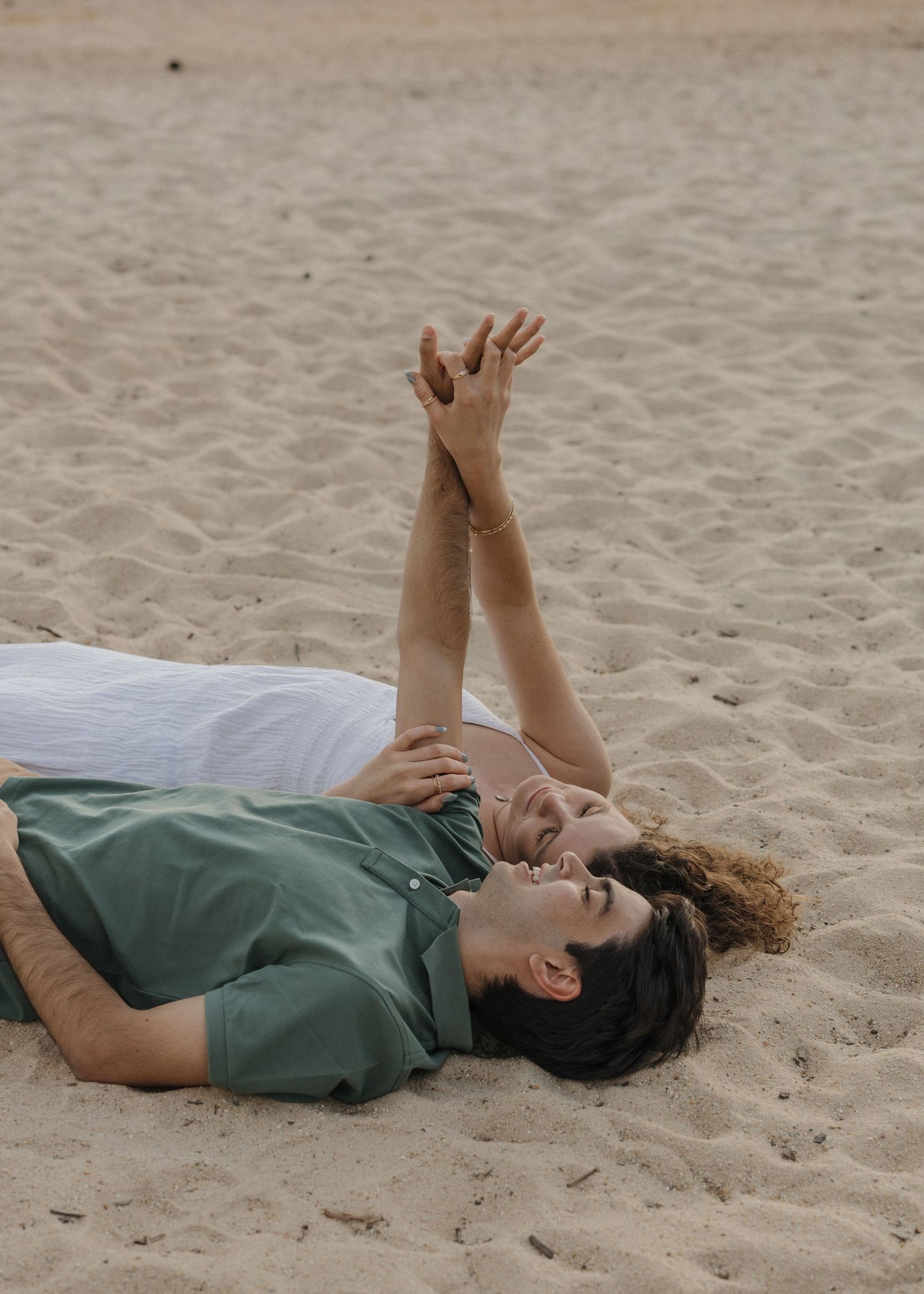 Couple lying on sandy beach holding hands raised, wearing green and white shirts, smiling romantically outdoors.
