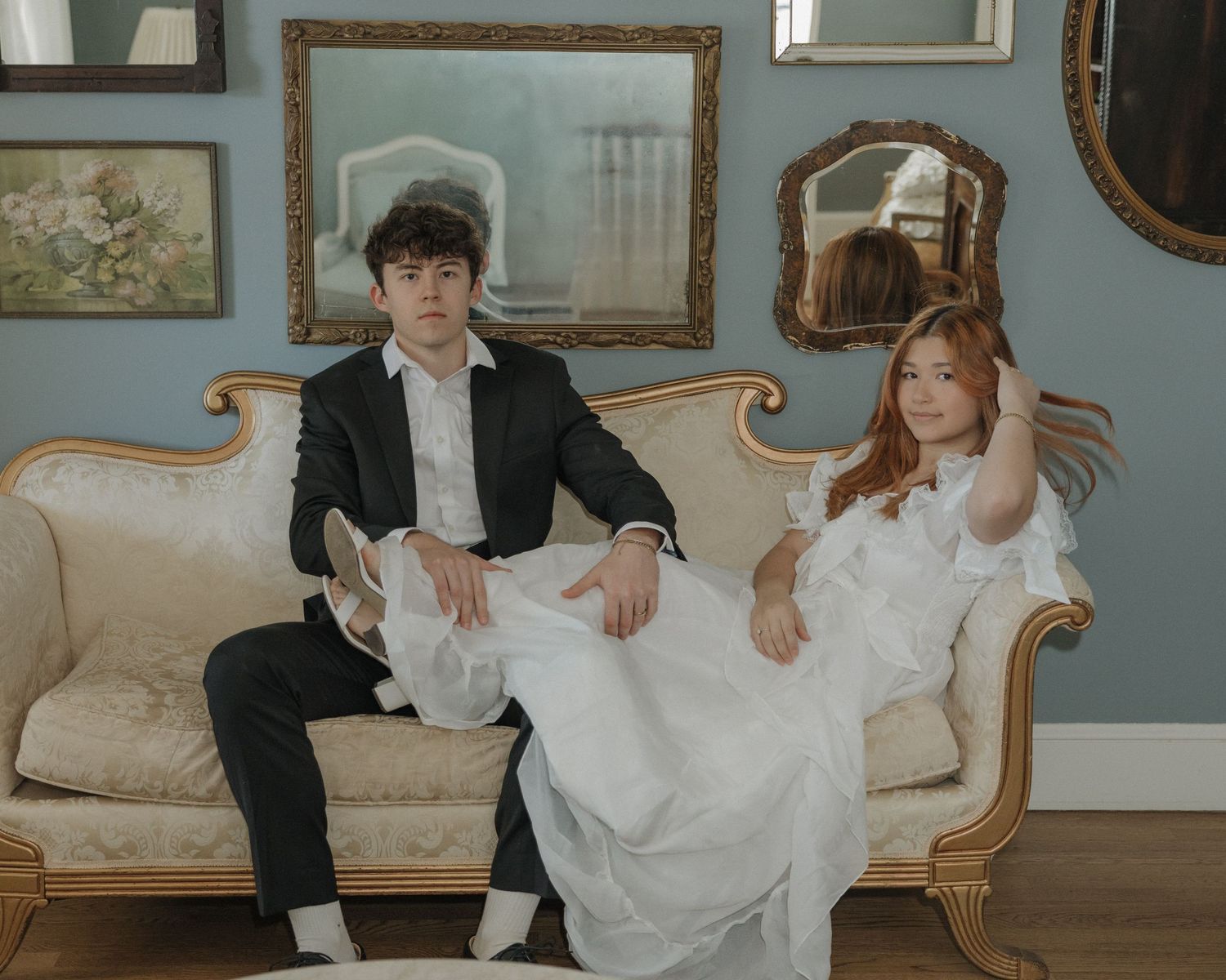 A couple in wedding attire sits together on an elegant vintage sofa in a room with ornate decor and blue walls.
