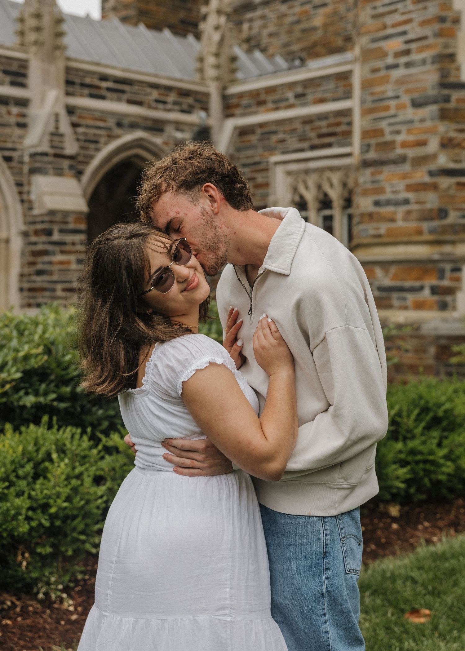 Couple embracing and kissing in front of a gothic stone building, woman in white dress wearing sunglasses.