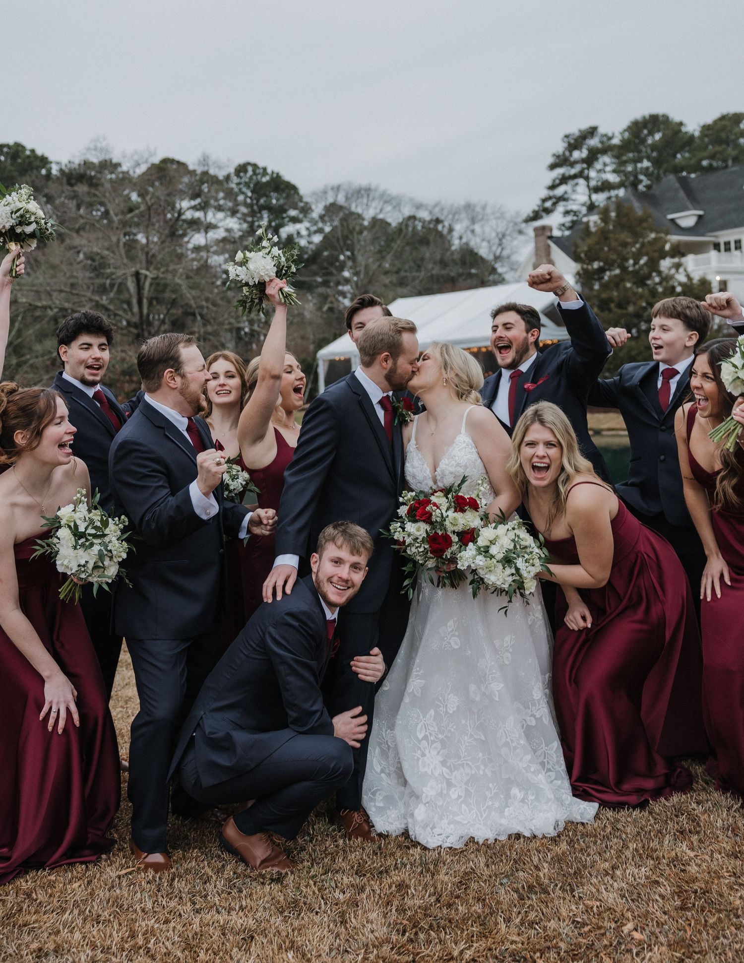 A joyful wedding party celebrates outdoors, bride and groom kissing, bridesmaids in burgundy gowns and groomsmen in navy suits.