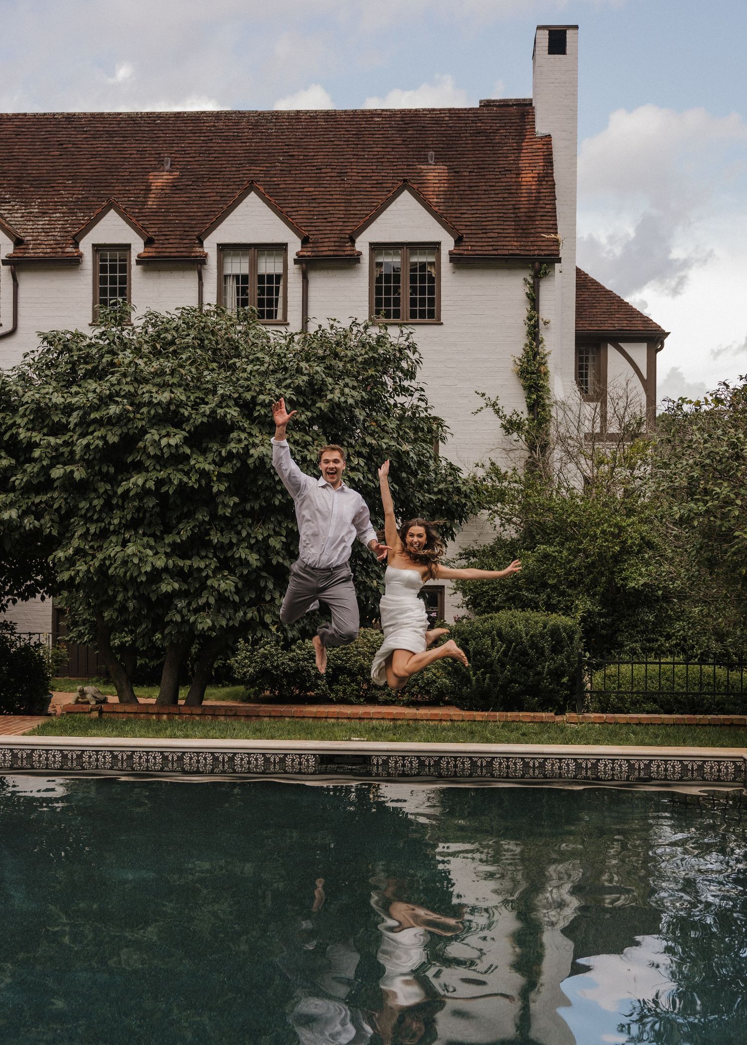 Joyful couple jumping with arms raised by a pool in front of an elegant Tudor-style mansion surrounded by lush greenery.