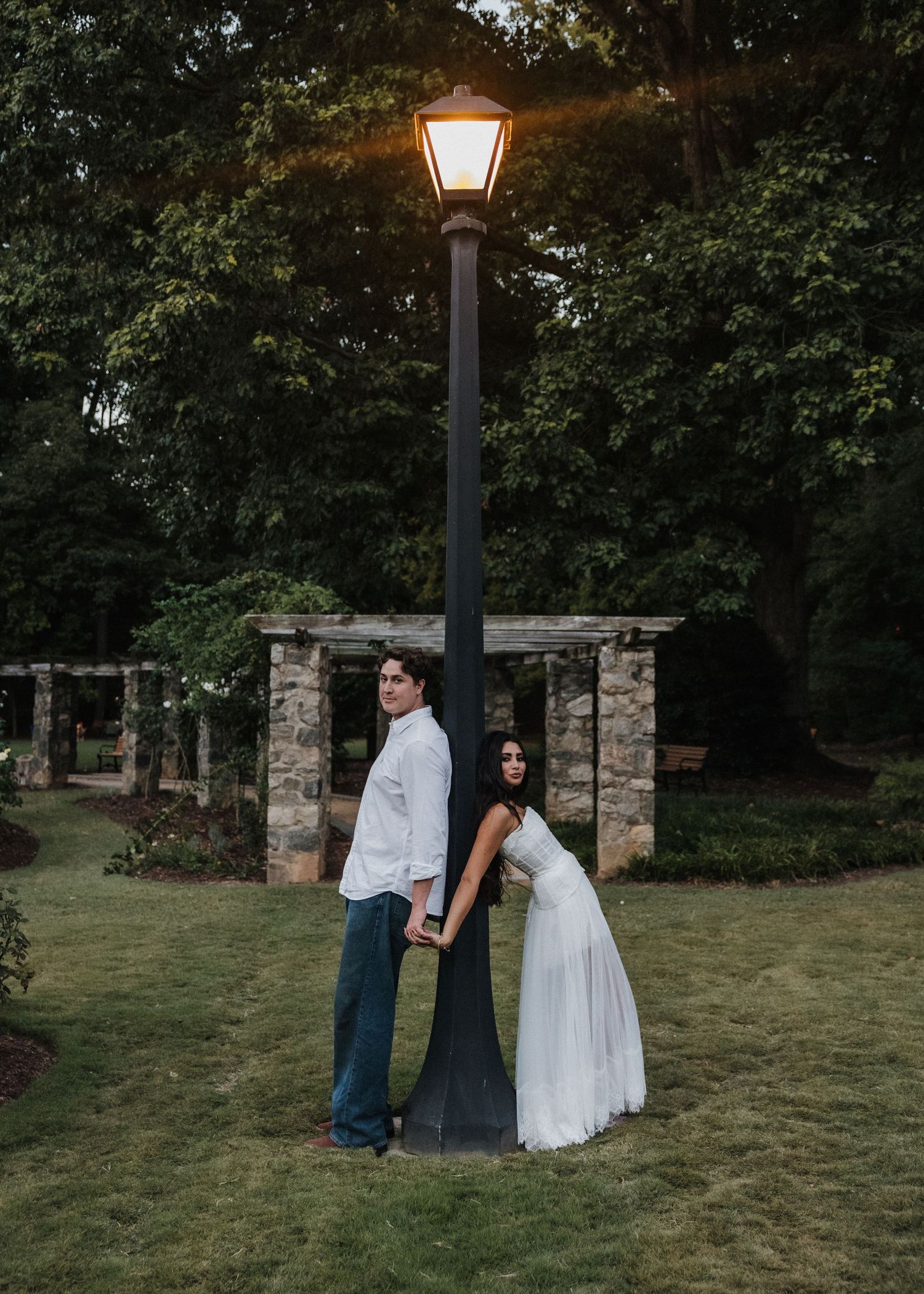 Couple holding hands back-to-back under a glowing street lamp in a lush garden at dusk.