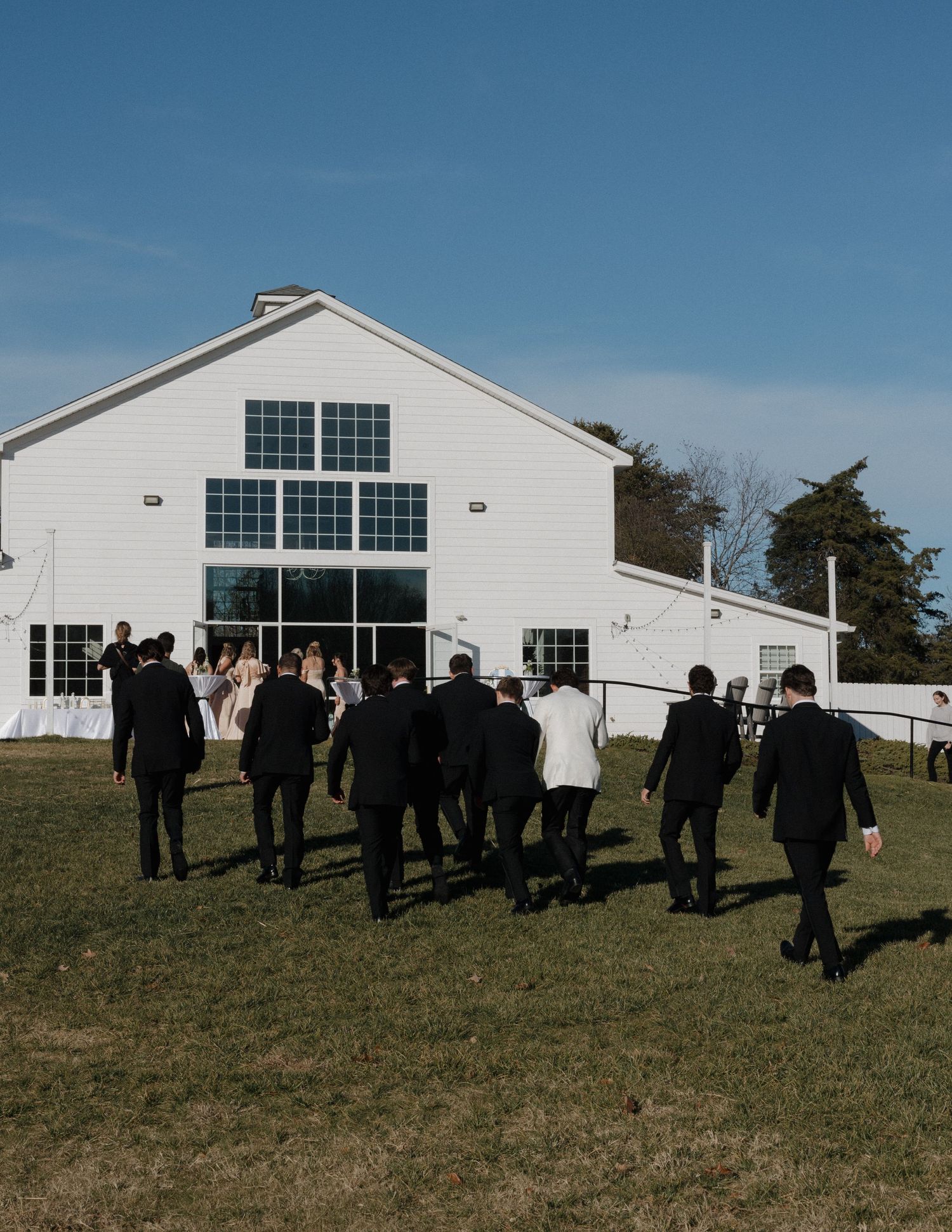 Wedding party walking toward a white barn venue on a sunny day with blue skies and green grass.