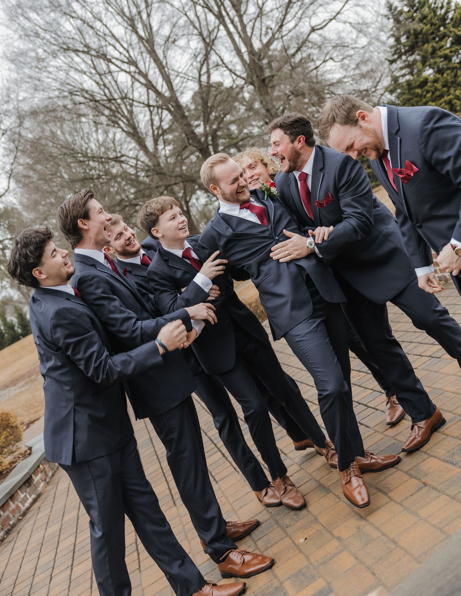 Groomsmen in navy suits and burgundy ties laughing and joking together outdoors at a wedding.