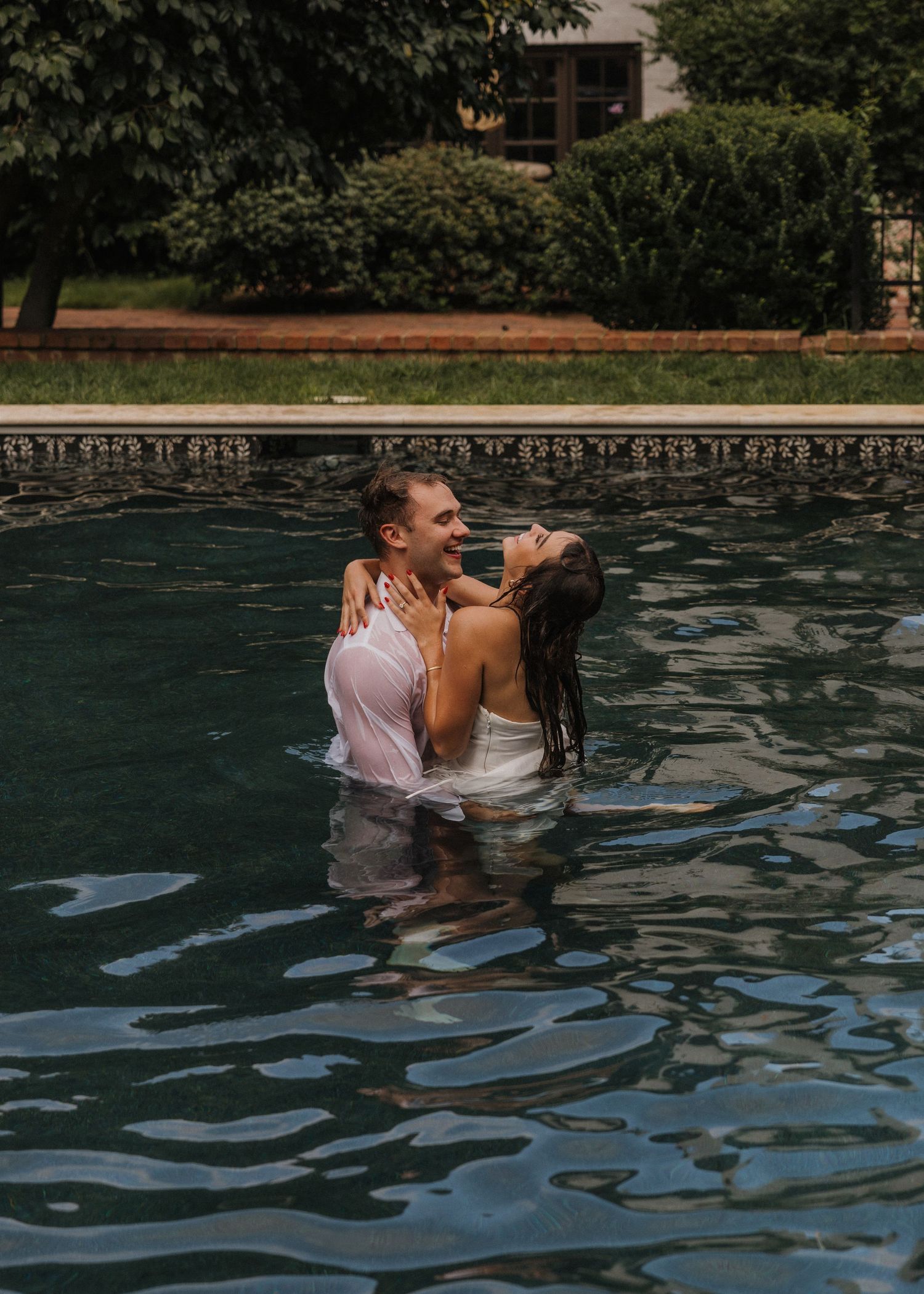 Couple embracing romantically in a pool, woman in white dress laughing joyfully, lush green trees in background.