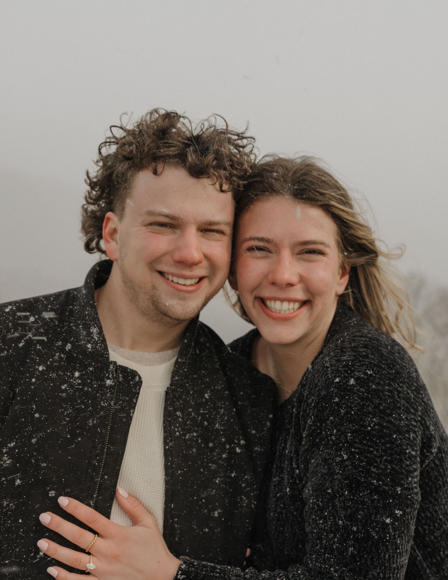 Happy couple smiling together outdoors in light snowfall, wearing dark jackets on a misty winter day.