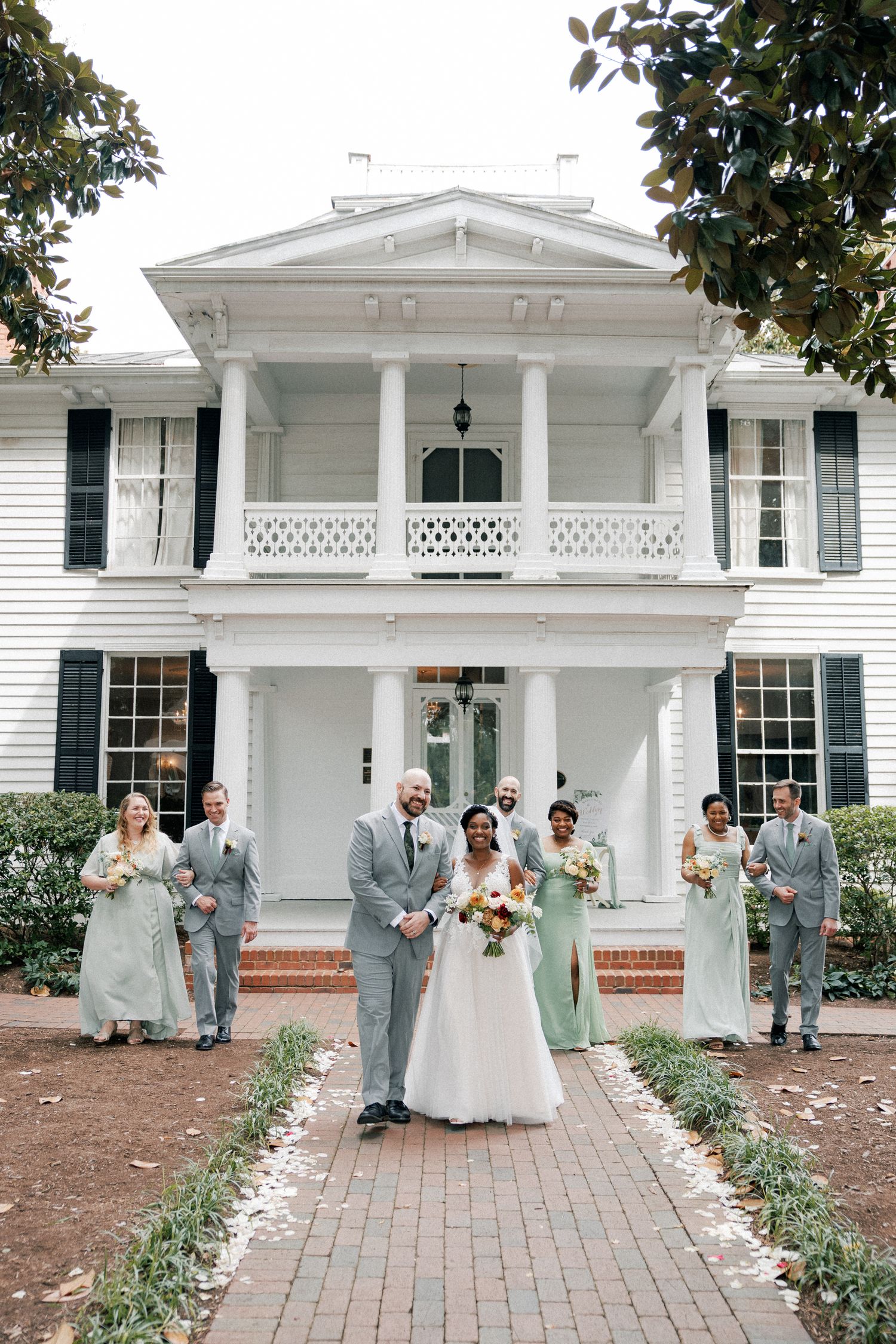 Wedding party in gray suits and white dress posing in front of elegant white colonial mansion.