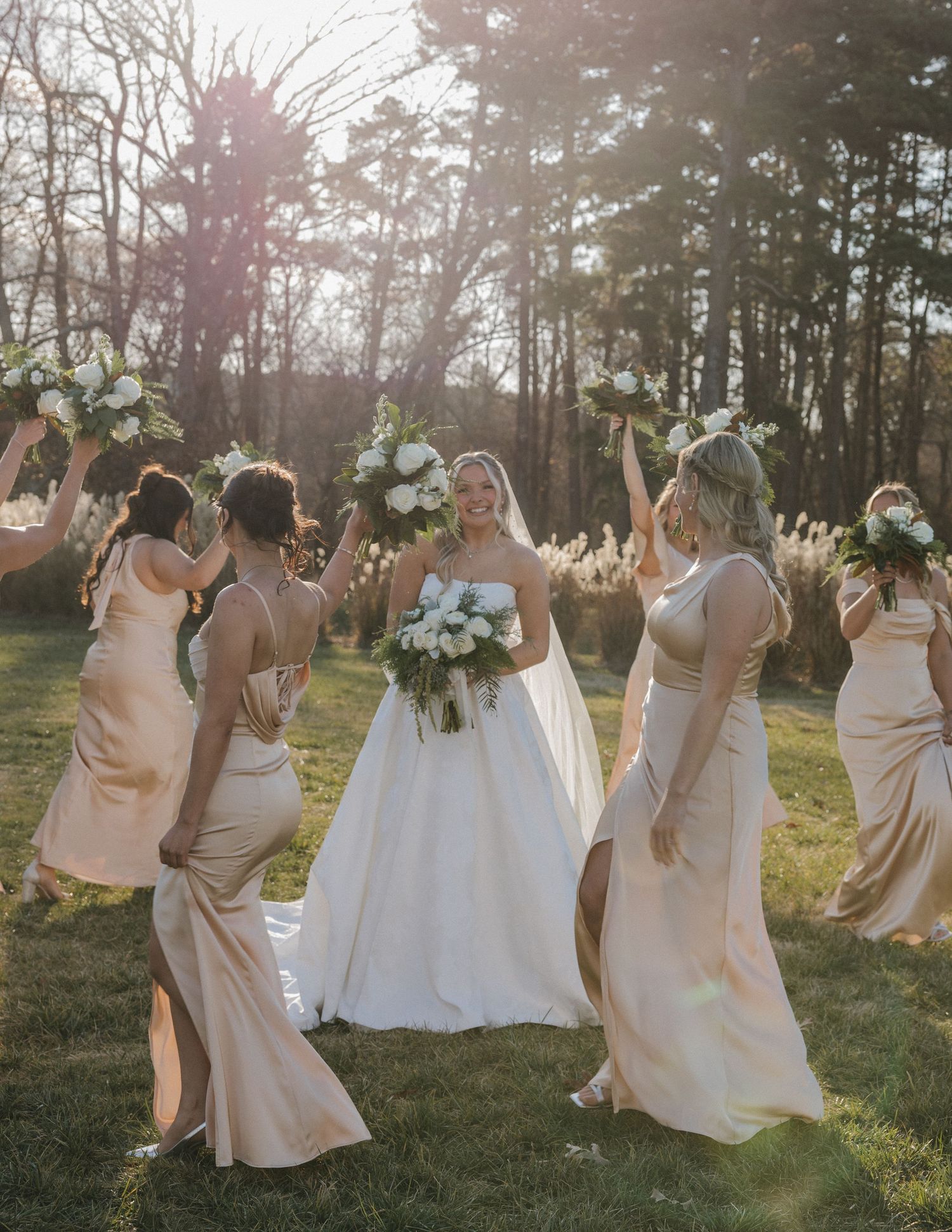 A bridal party in champagne colored dresses dances joyfully together in a sunlit field at golden hour.