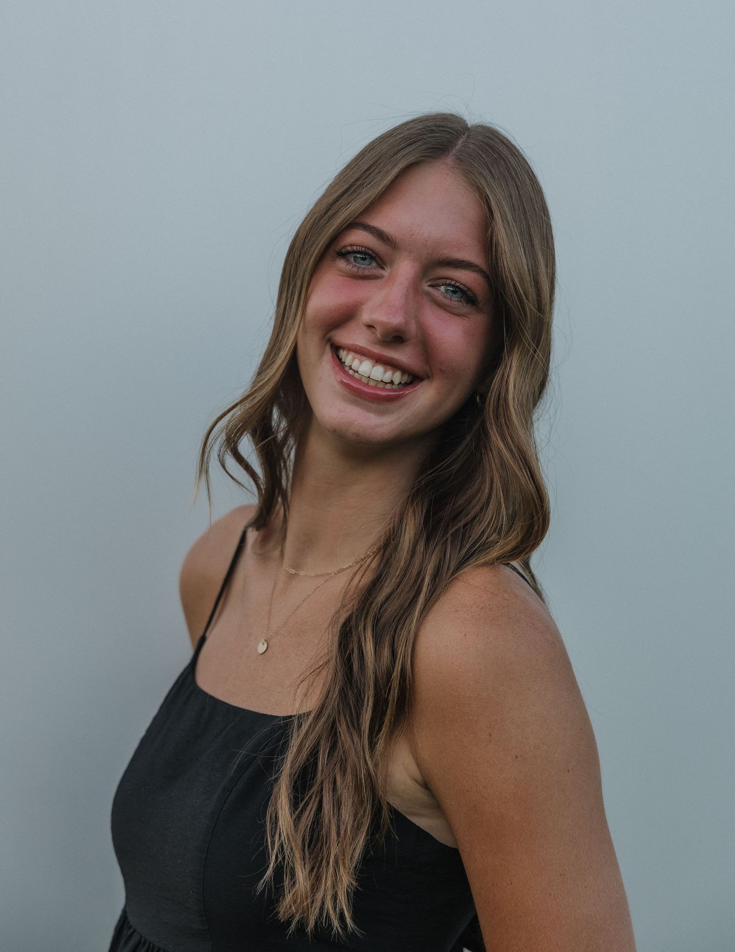 Smiling young woman with long brown hair wearing a black top and delicate necklace against a gray background.
