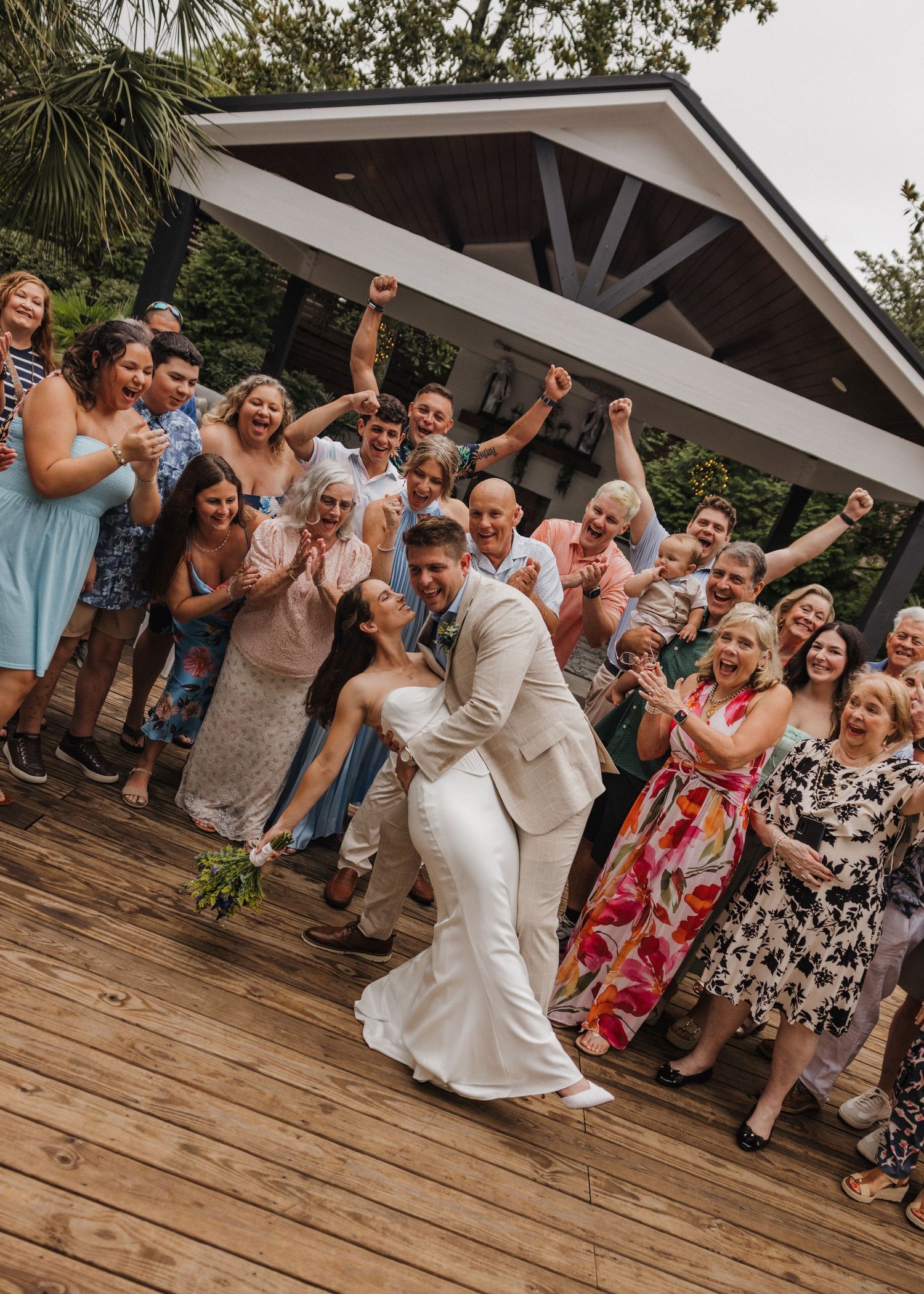 Joyful wedding couple shares a romantic dip kiss surrounded by cheering guests on a wooden deck.