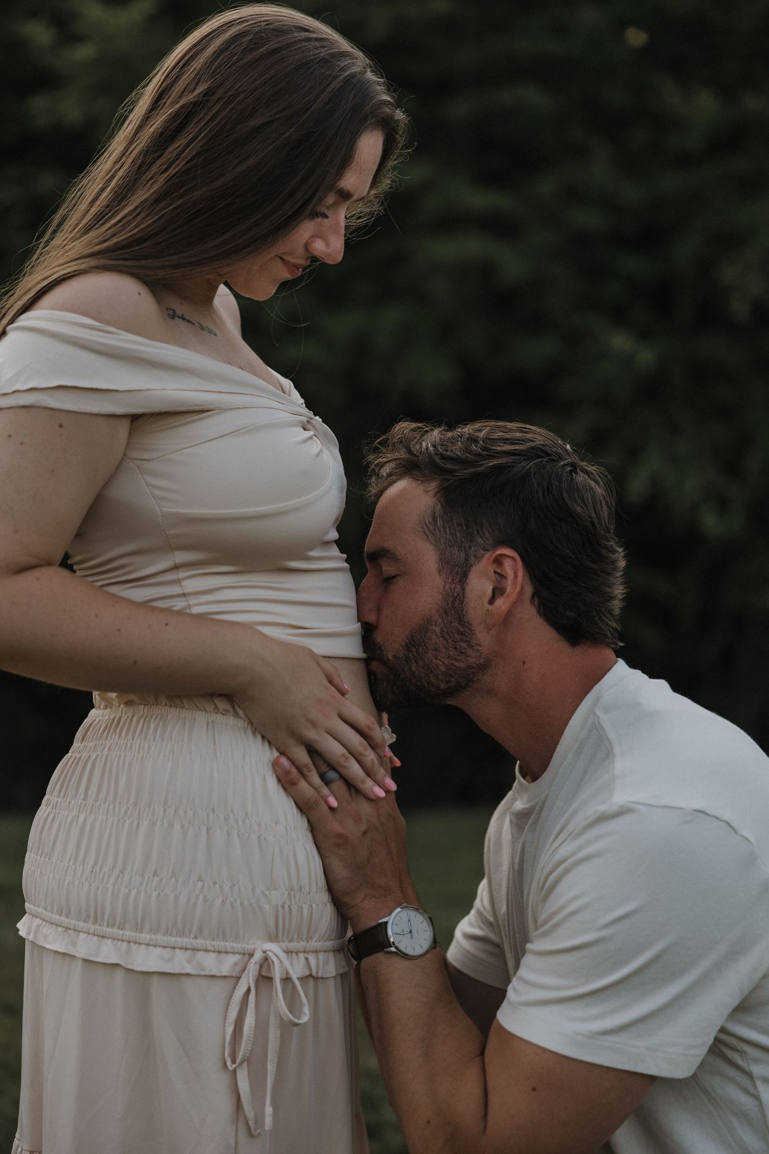 A man kneeling and kissing a pregnant woman's belly outdoors during a maternity photoshoot.