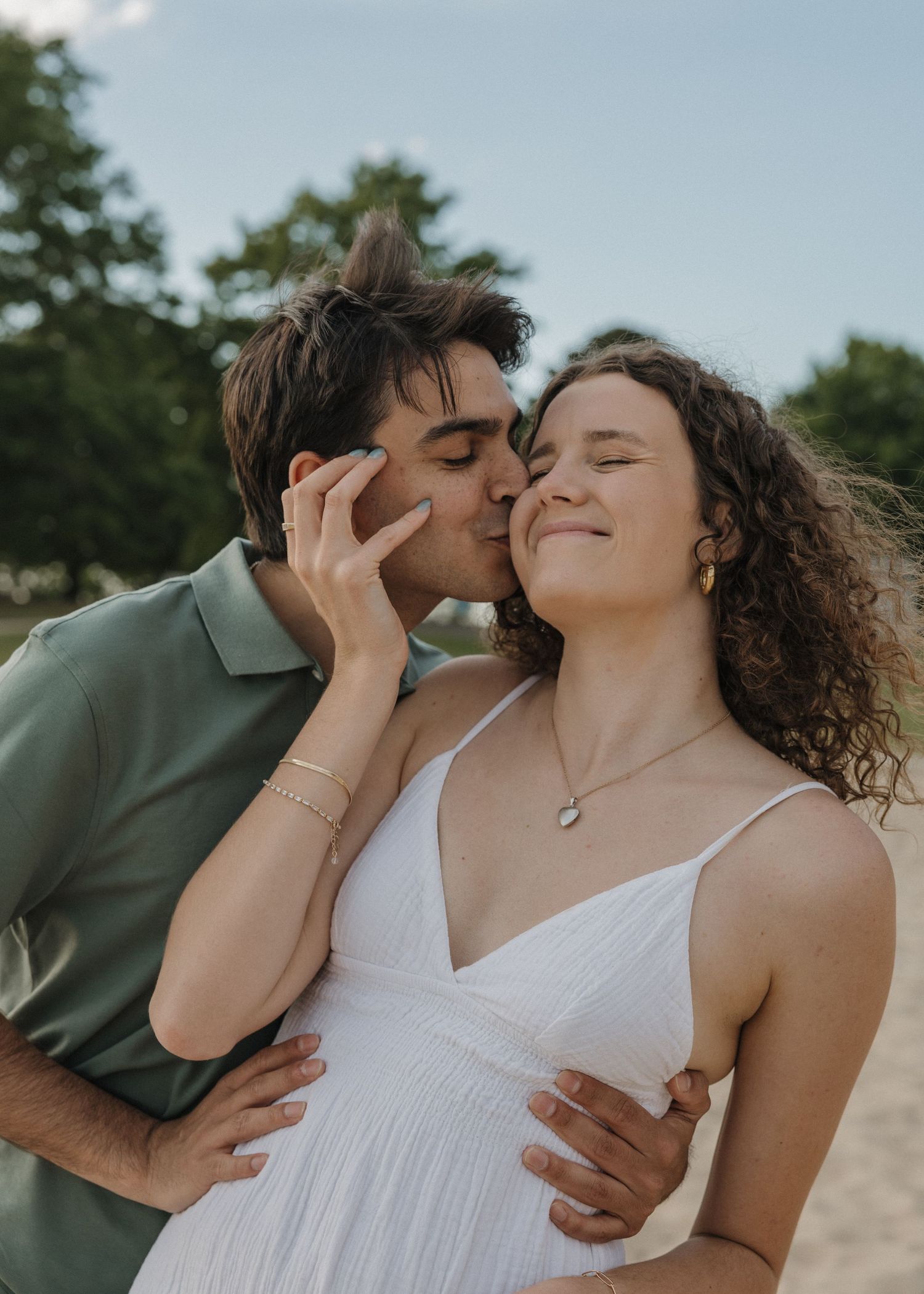 A young couple shares a tender moment outdoors, man kissing womans cheek as she smiles in a white dress.