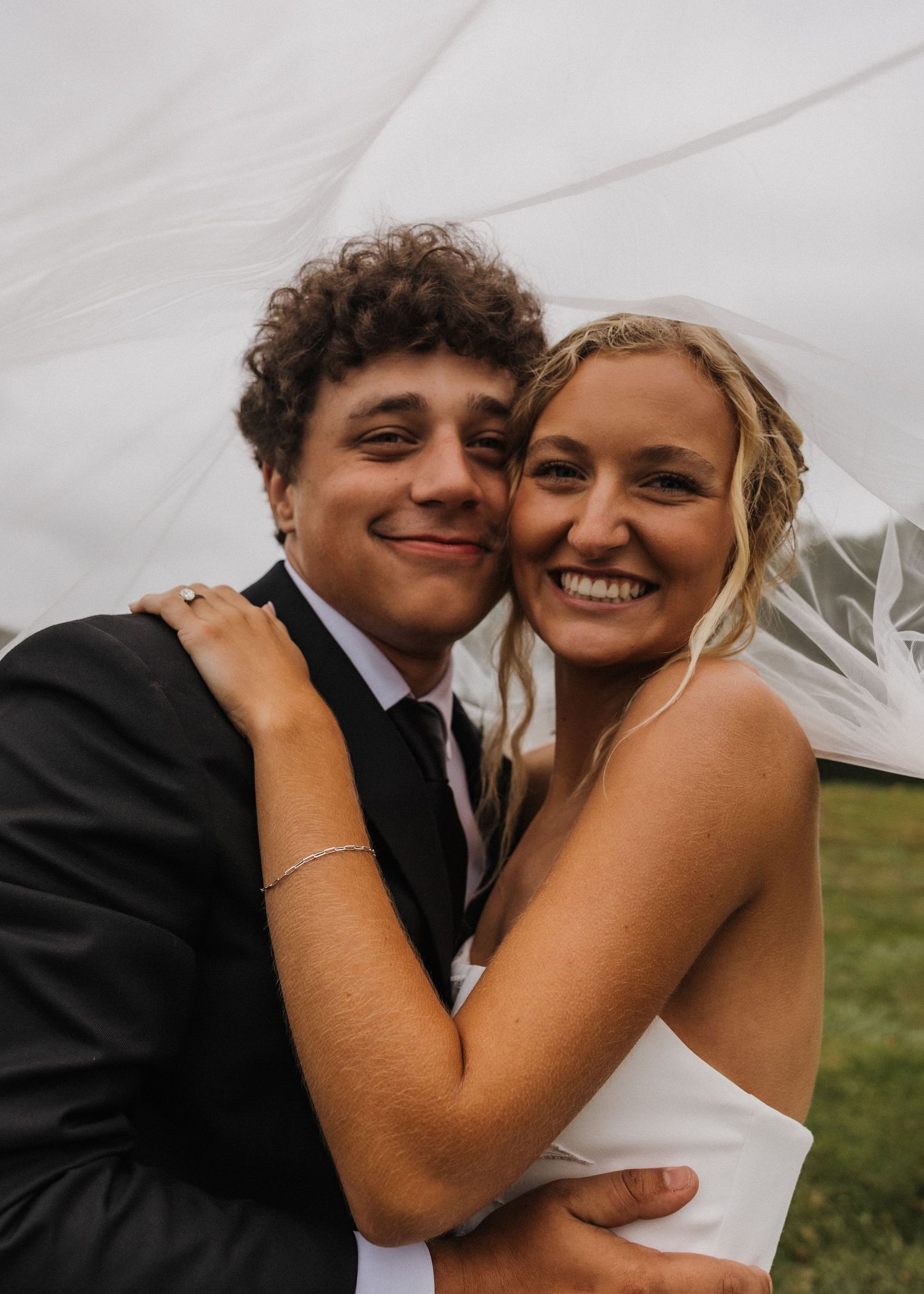 Bride and groom smiling under wedding veil, groom in black suit, bride in white dress, outdoor wedding photo.