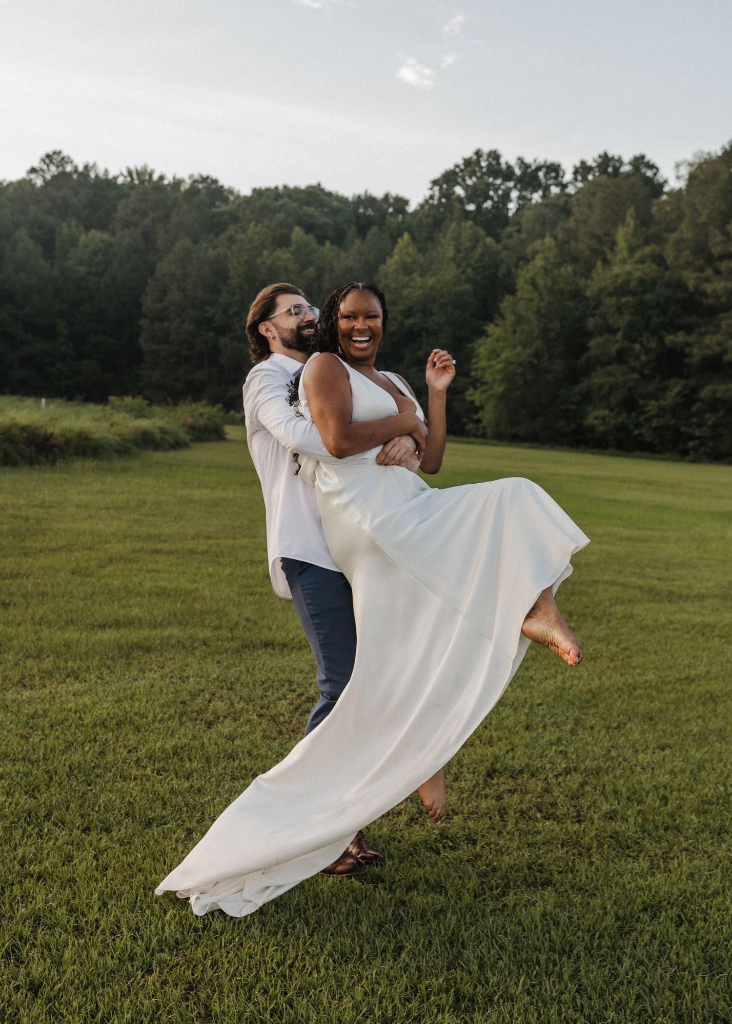 Groom lifting smiling bride in white dress outdoors on green lawn with trees in background.