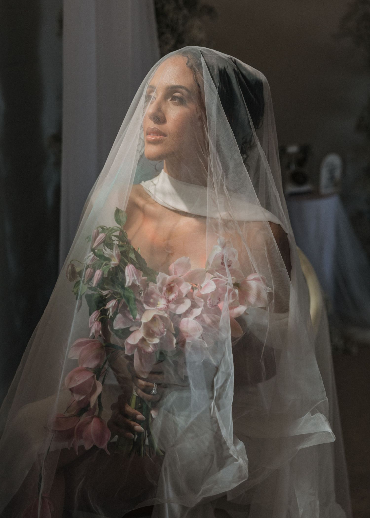 Elegant bride wearing a sheer veil holding pink floral bouquet in a softly lit room.