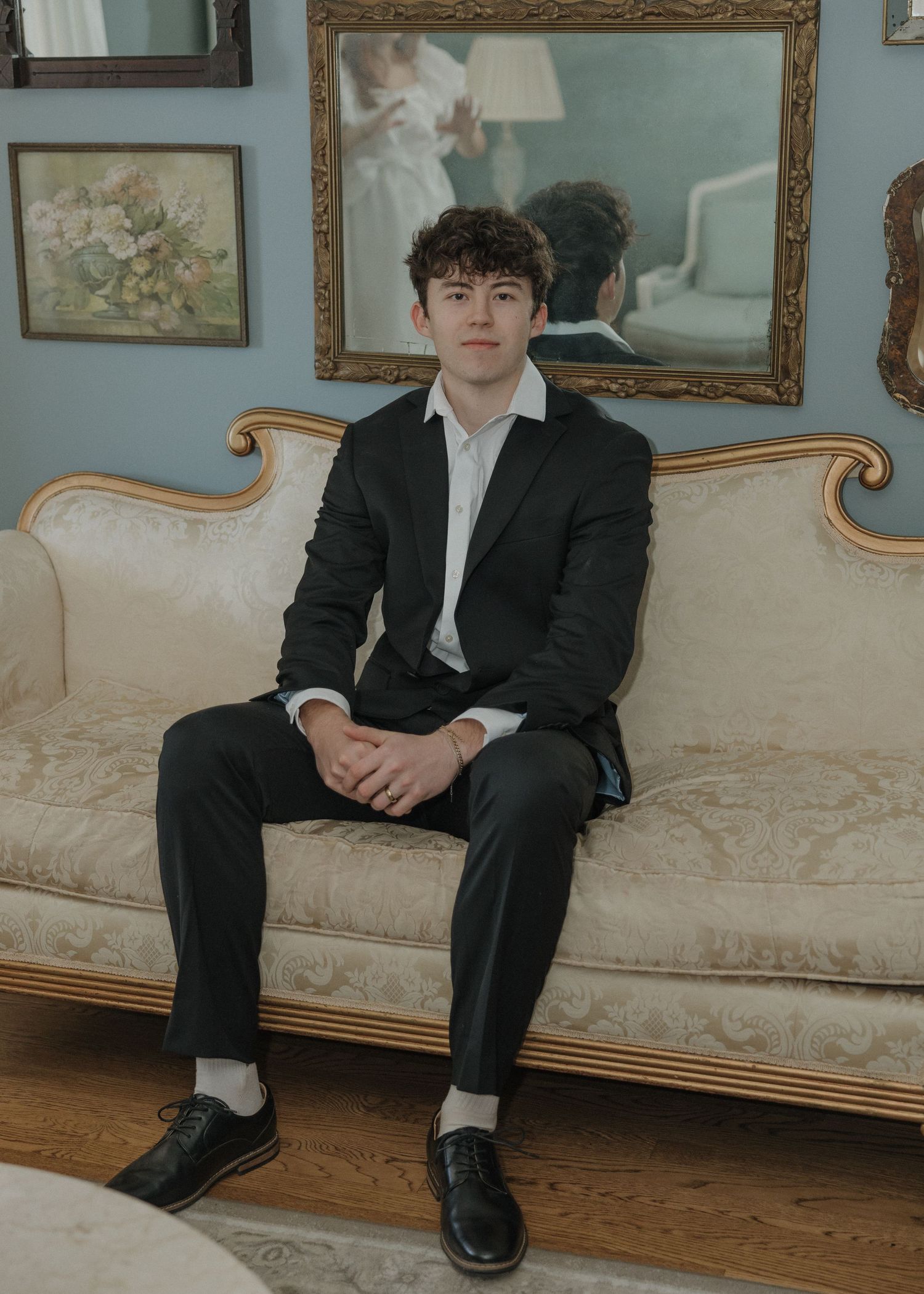 A person in a black suit sits on an elegant cream-colored vintage sofa with ornate framed artwork visible on the wall behind.