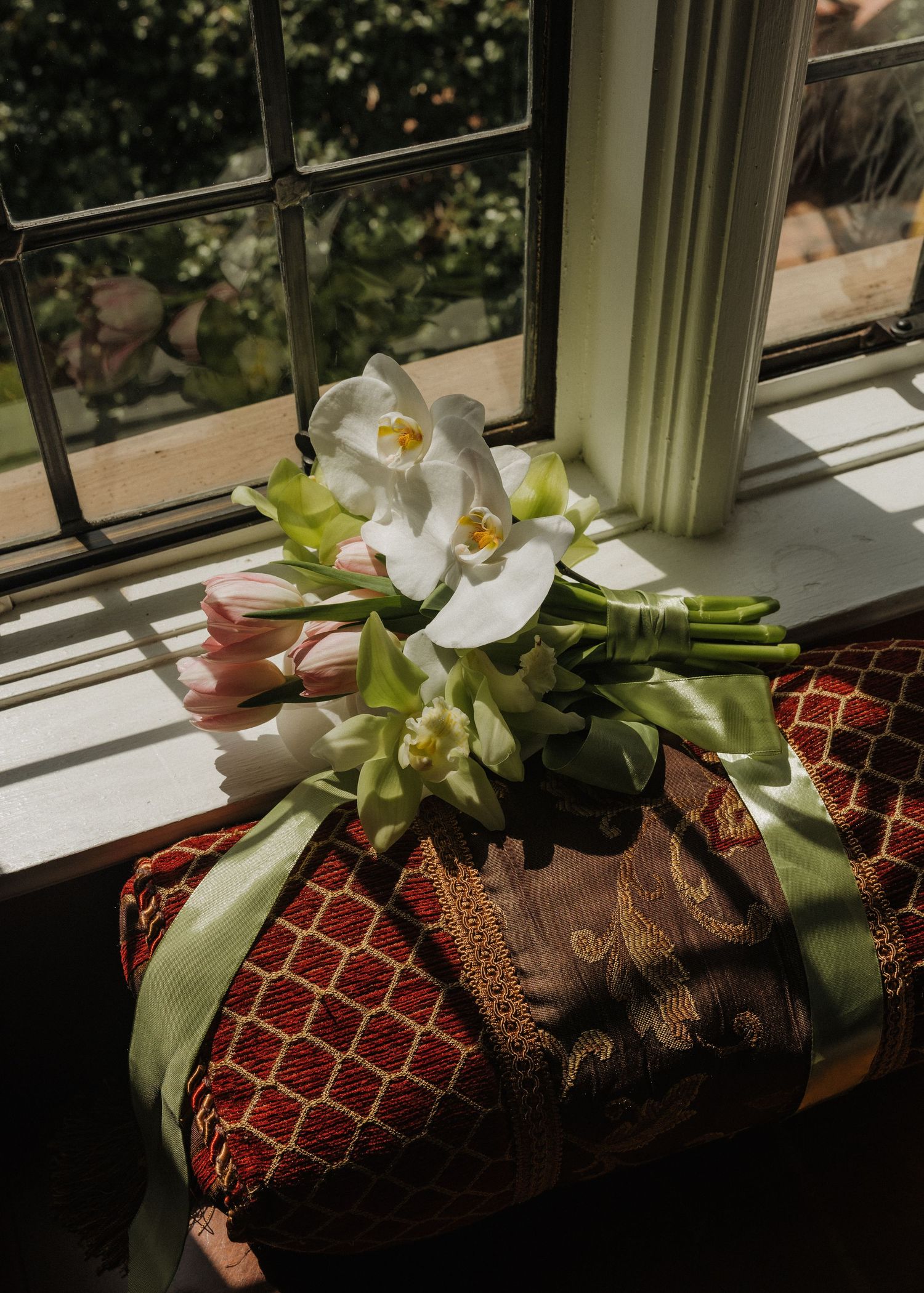White orchids and pink tulips bouquet tied with green ribbon resting on decorative red cushions near a sunlit window.