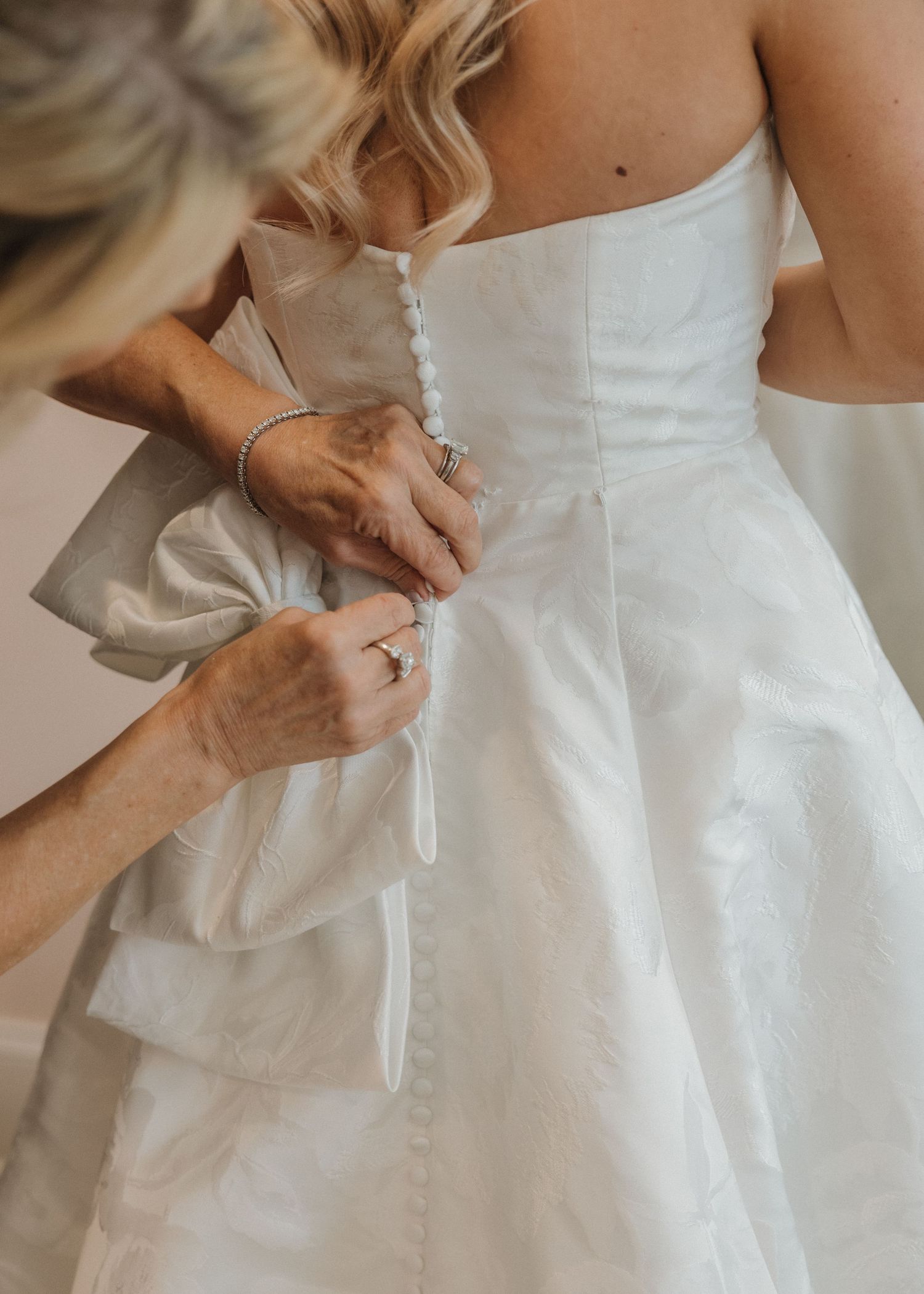 A helper fastens buttons on the back of a white strapless wedding gown with a large bow detail.