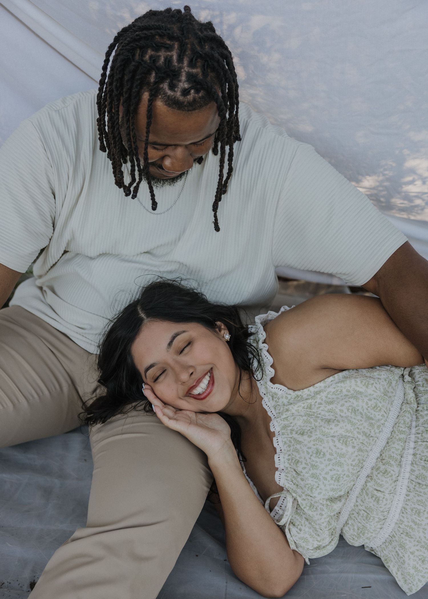 A smiling woman rests her head on a man's lap as they relax together on a blanket in a cozy, intimate setting.