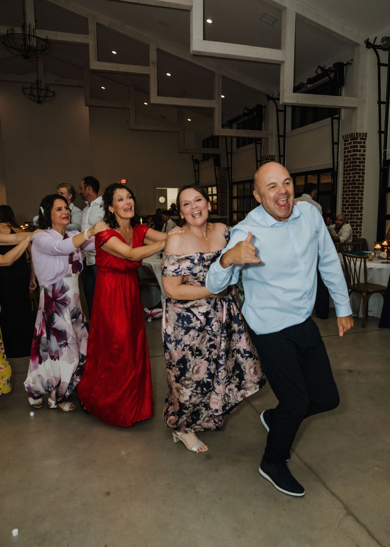 Guests dancing joyfully at a wedding reception in an elegant venue, with women in formal gowns and a man in a light blue shirt.