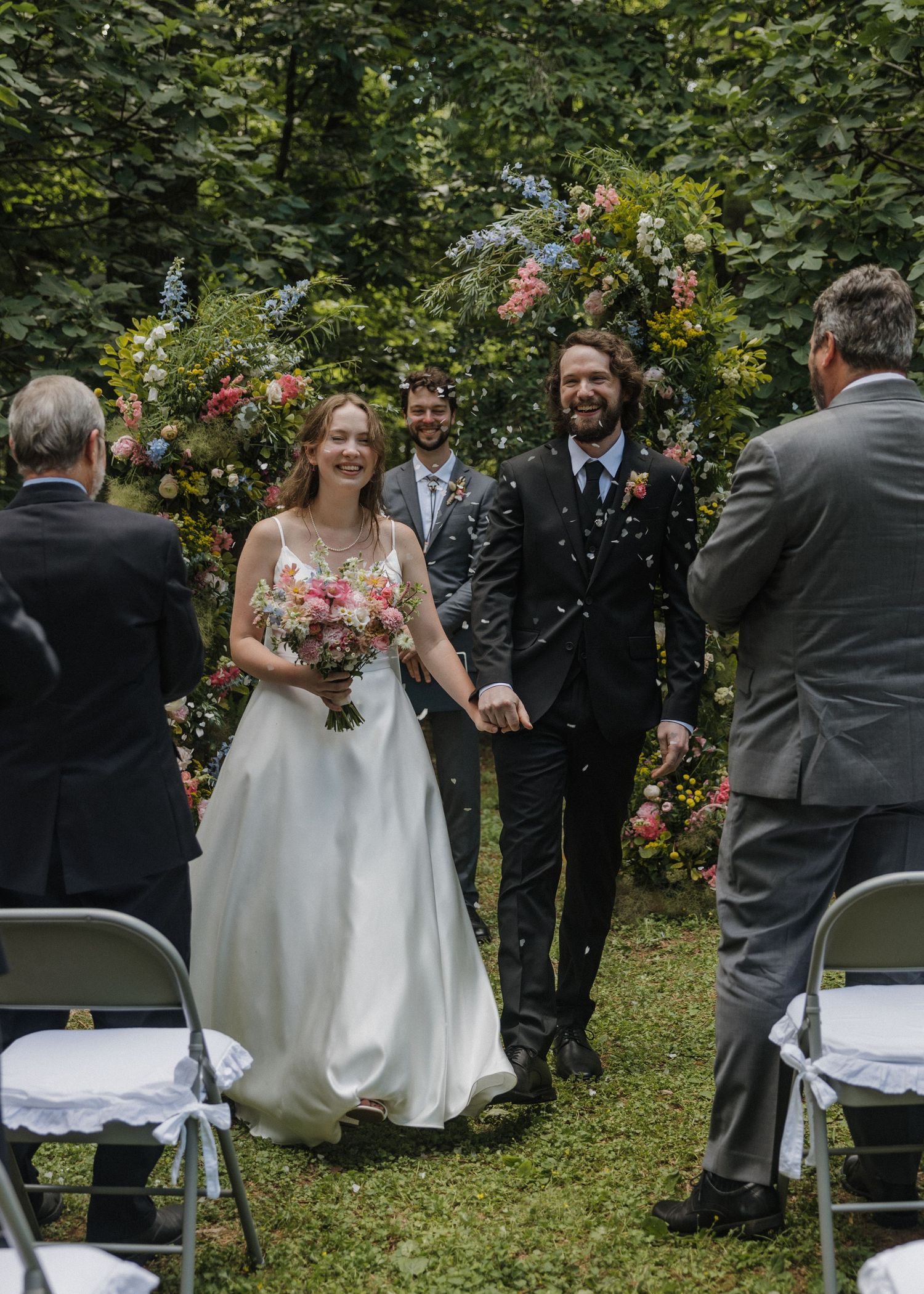 Bride and groom walk down outdoor aisle after wedding ceremony, surrounded by lush greenery and a floral arch.